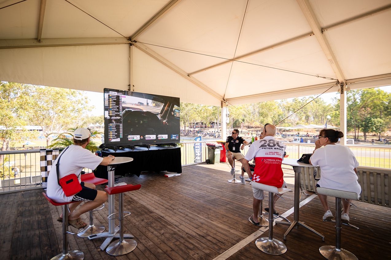 People sitting under a tent watching a large screen displaying a race, with trees visible in the background.