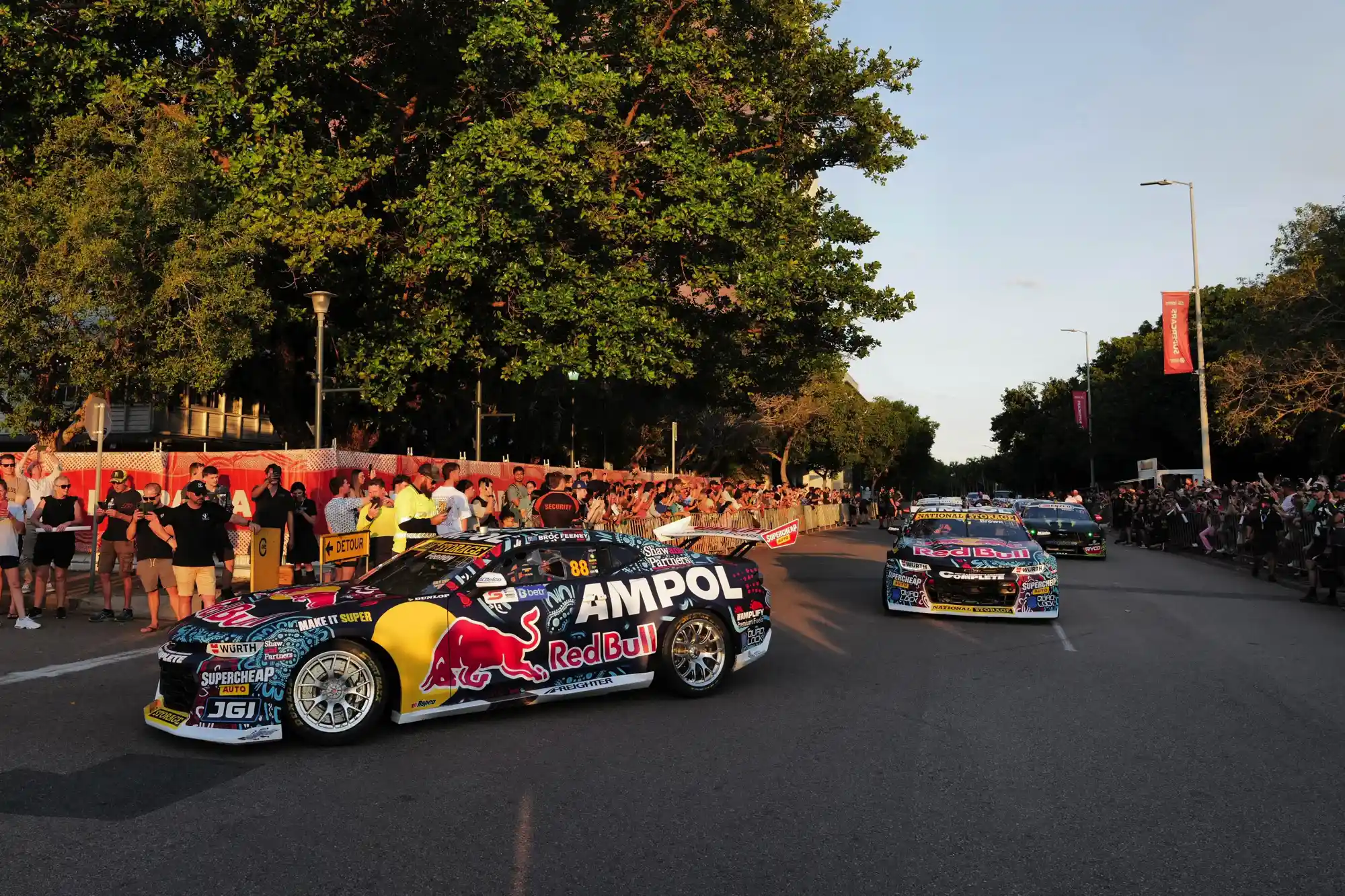 Race cars with Red Bull and Ampol branding speed down a street lined with spectators and trees during a motorsport event.
