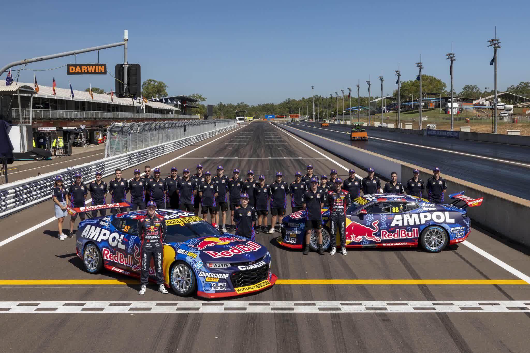 Race cars with Red Bull and Ampol branding on a track, surrounded by a large team in matching attire. A sign reads "Darwin" in the background.