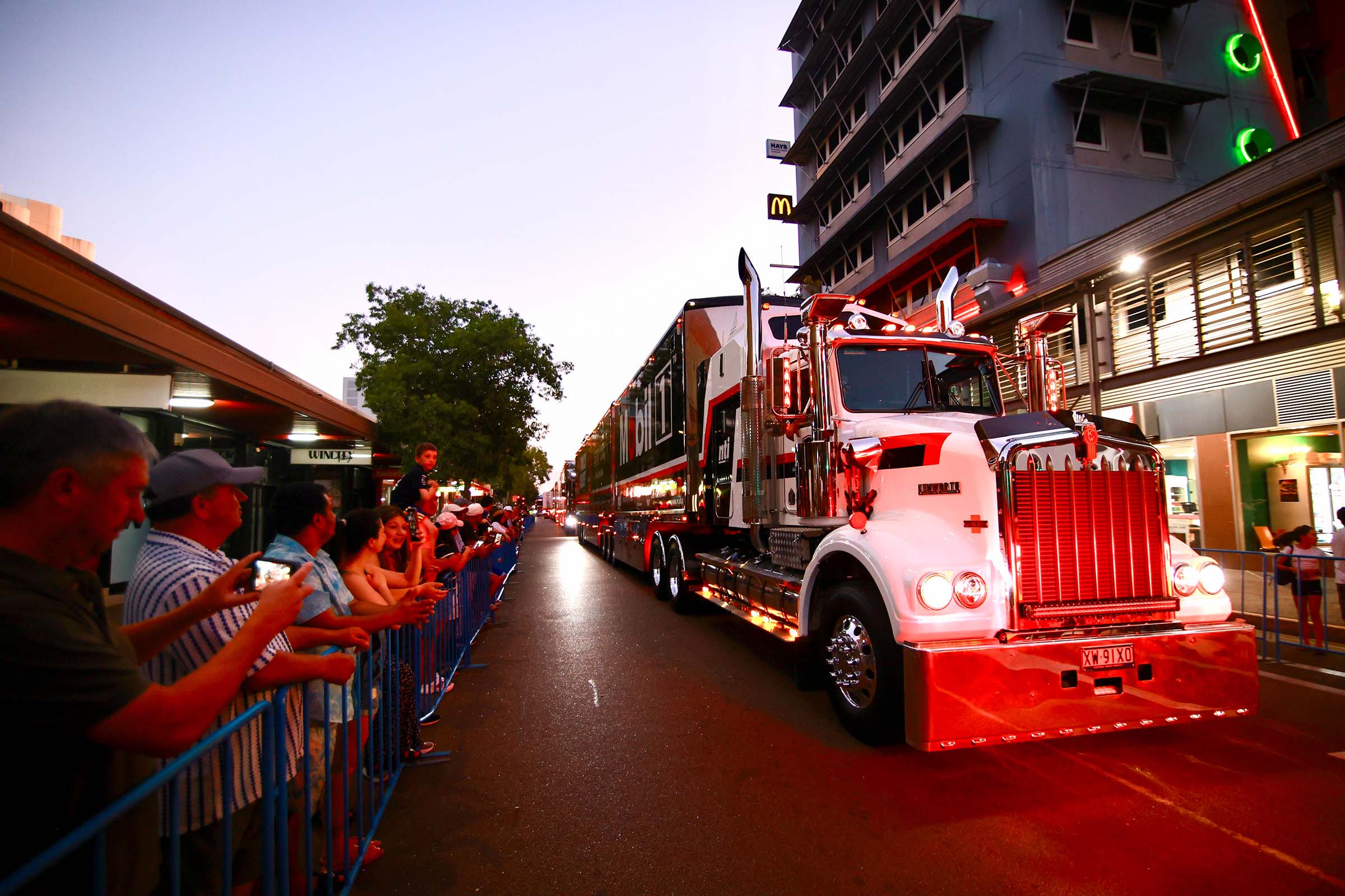 A large white Supercars transporter truck passing through Darwin city at dusk with spectators lining the street.
