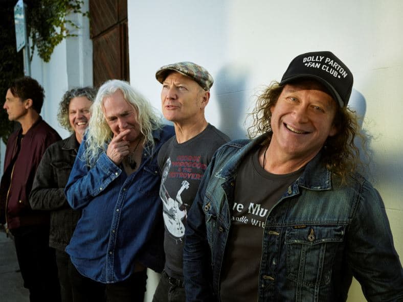 Five men stand outdoors, smiling and casually dressed, with one wearing a "Dolly Parton Fan Club" hat.