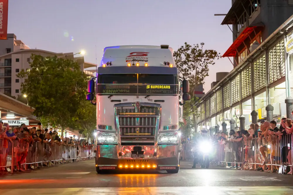 A large truck with bright lights drives down a city street at dusk, surrounded by crowds behind barriers, under a clear sky.