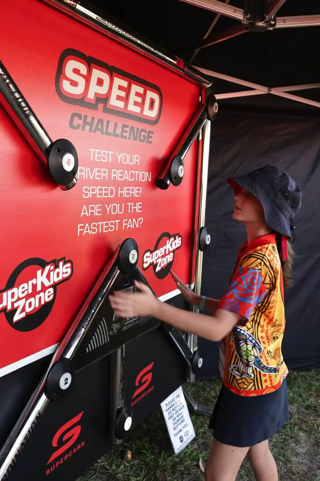 A person in a colorful shirt and hat plays a reaction speed game at a "SuperKids Zone" booth, pressing buttons on a red board.