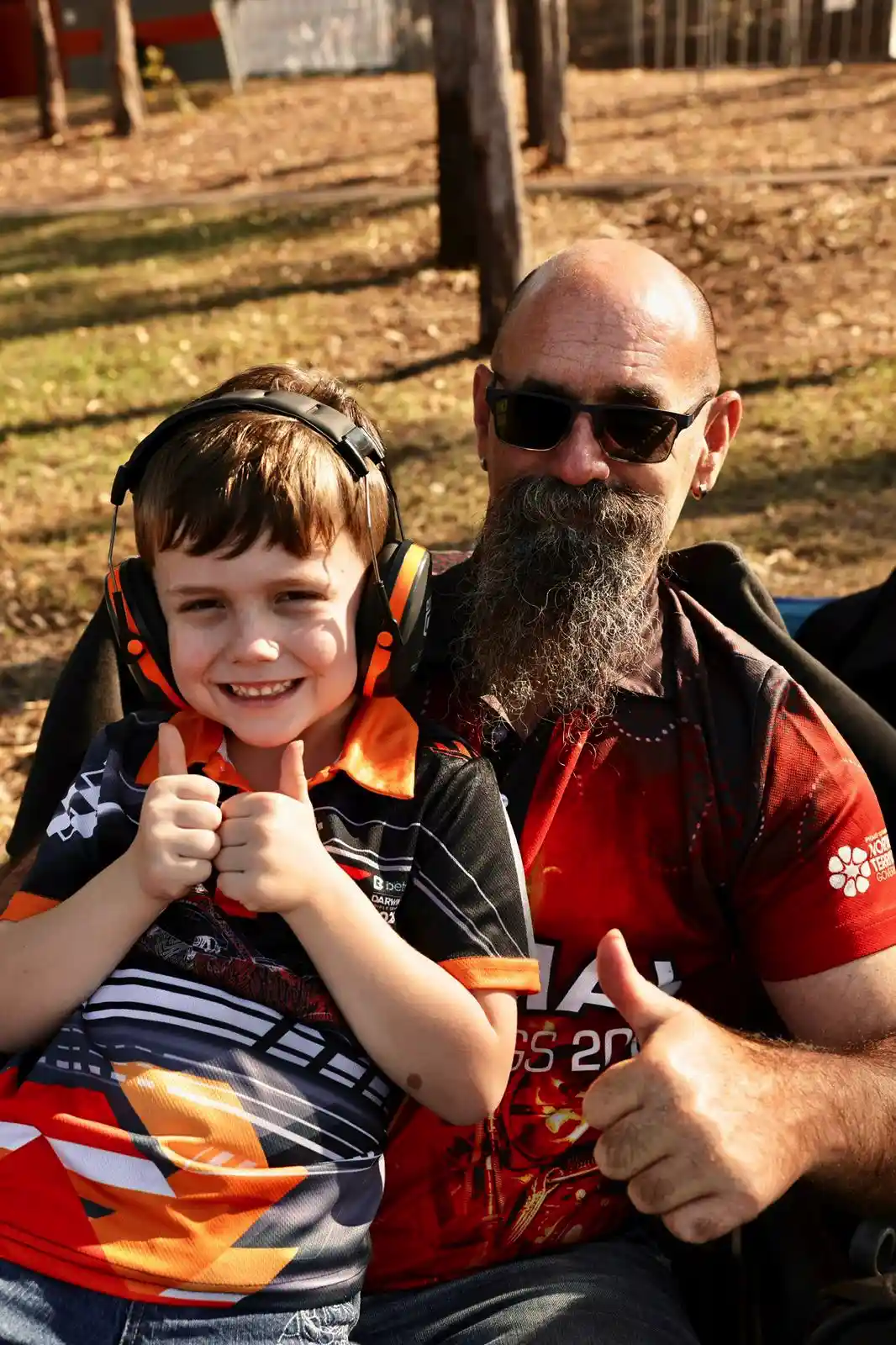 A bearded man and a smiling child with headphones give thumbs up outdoors, sitting together in a sunny park.