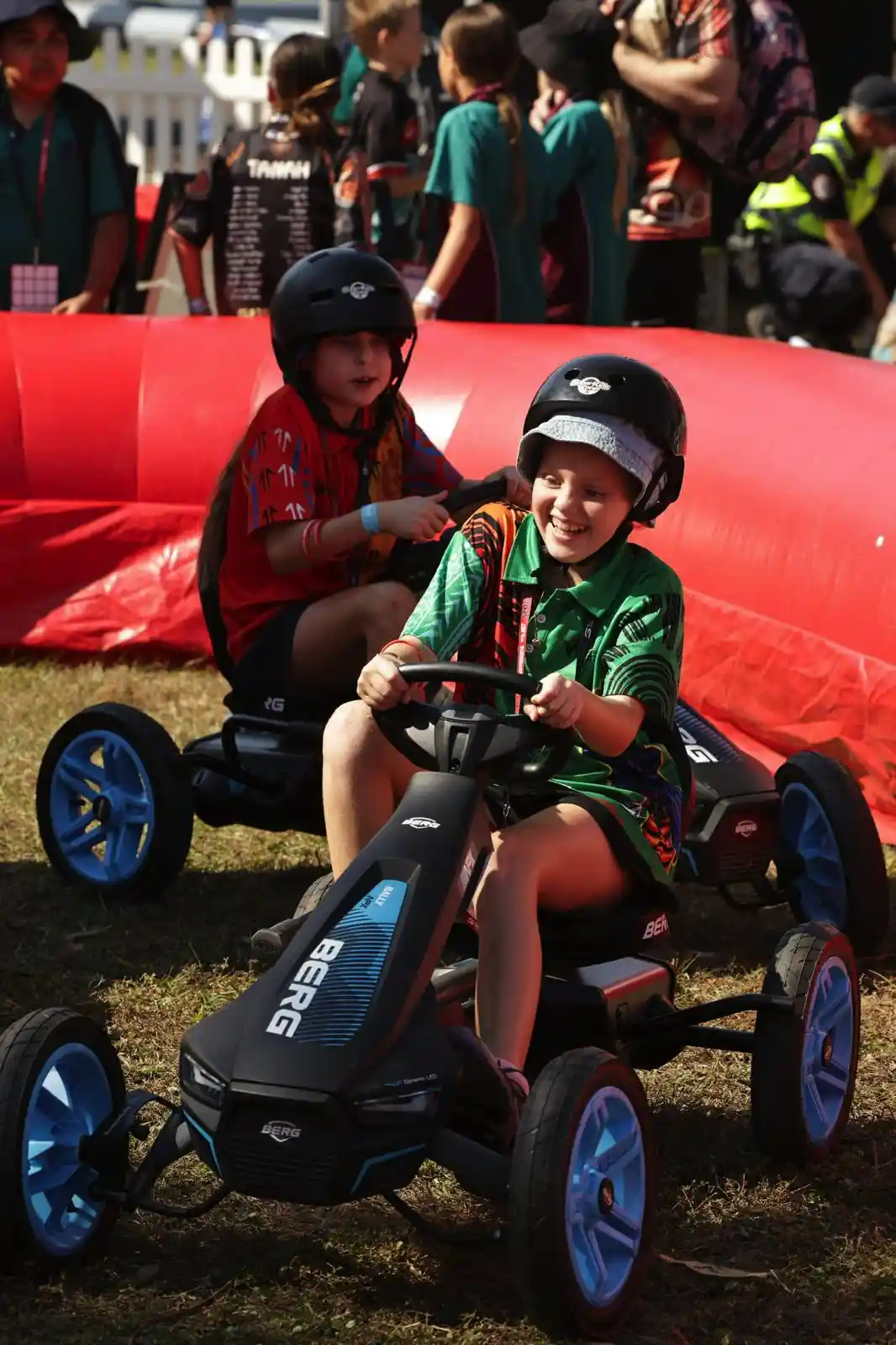 Two children wearing helmets race pedal karts on grass, surrounded by a red inflatable barrier, with spectators in the background.