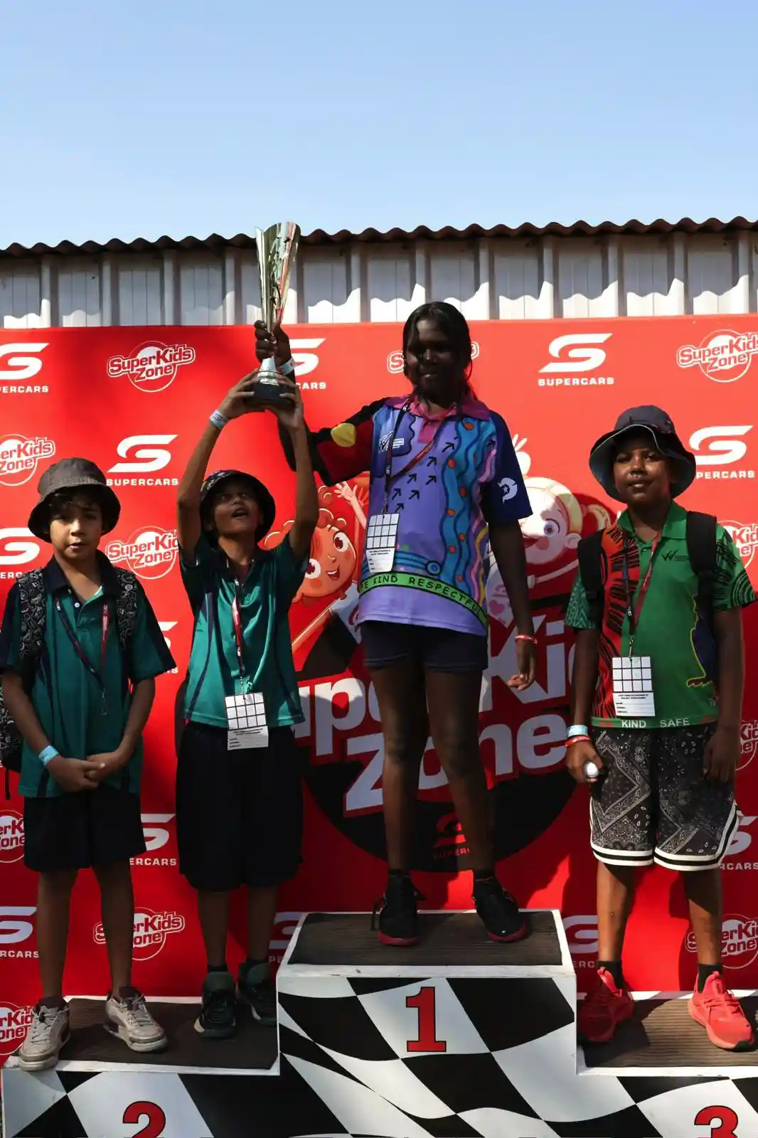 Four children stand on a podium with a trophy. The backdrop features a red "Supercars" logo. The child on the top step holds the trophy.