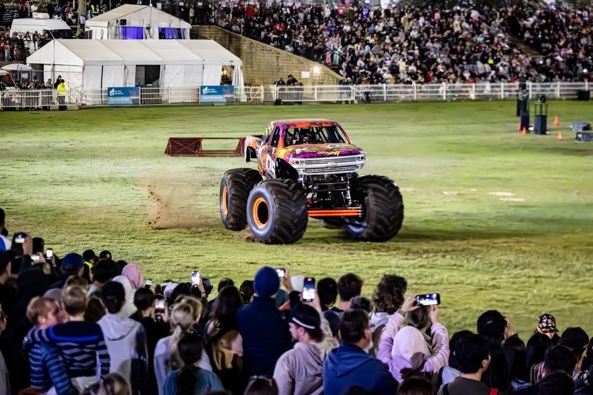 A purple and orange monster truck tears up a grassy field as a crowd watches, many filming, with stadium seats behind.