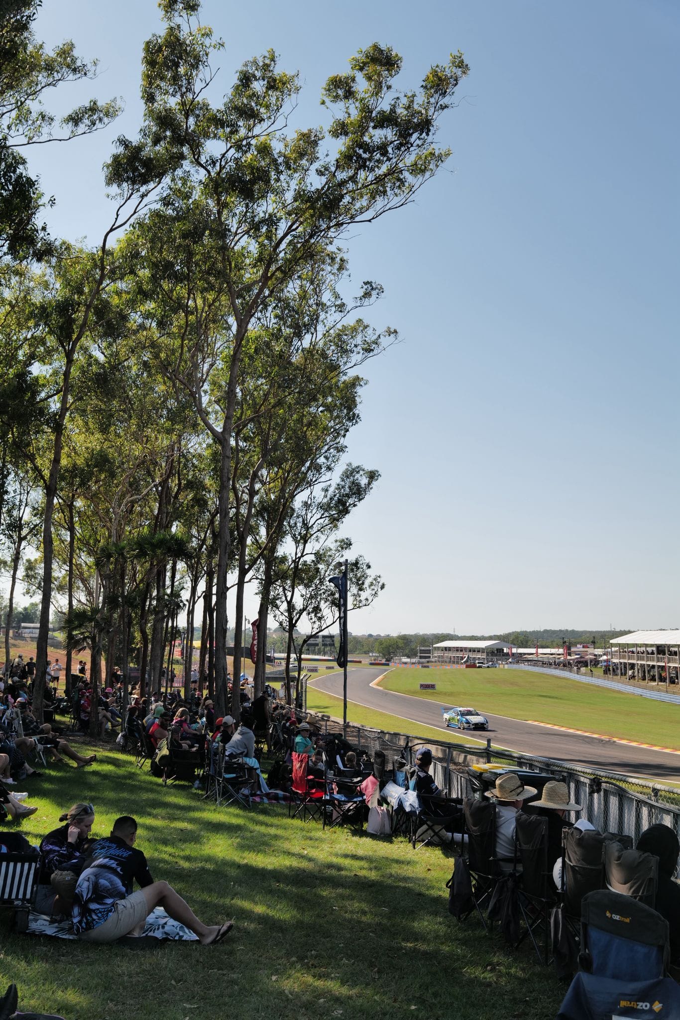 Spectators watch a Supercar race around Hidden Valley Raceway from a shaded grassy viewing area lined with trees.