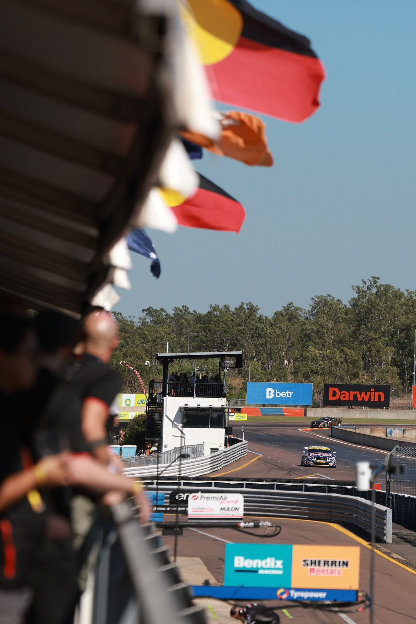 Supercars race past the pit building at Hidden Valley Raceway, with Aboriginal flags flying above the garages.