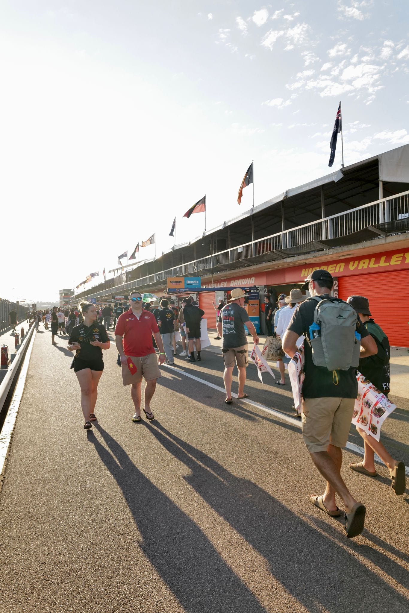 Fans walk along pit lane at Hidden Valley Raceway during Darwin Supercars, with team garages and flags lining the circuit in warm morning light.