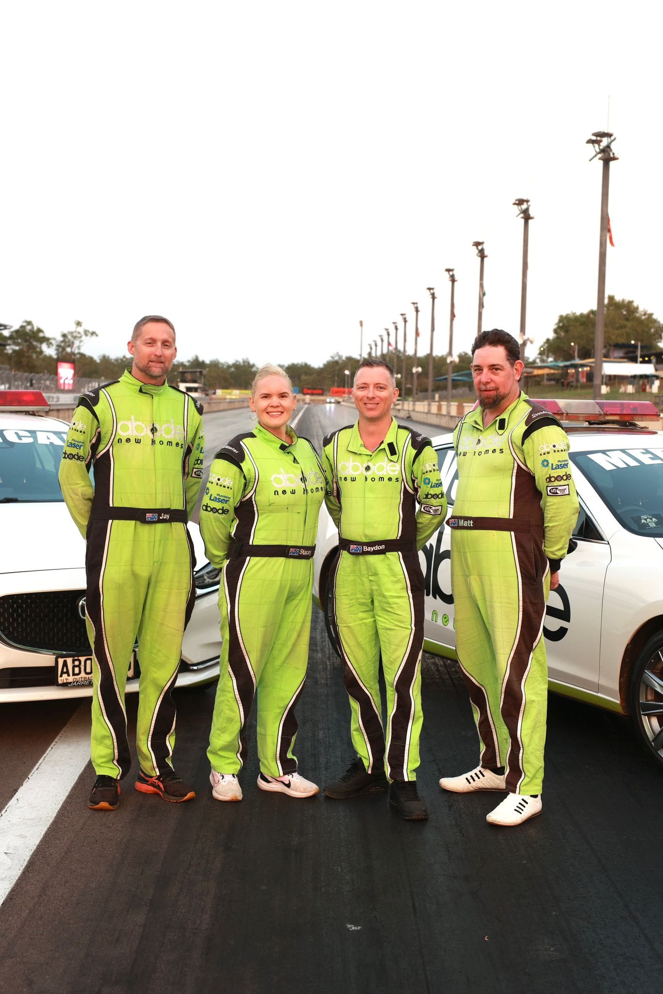 Four track safety crew members in green racing suits pose beside official vehicles on the Hidden Valley Raceway main straight.