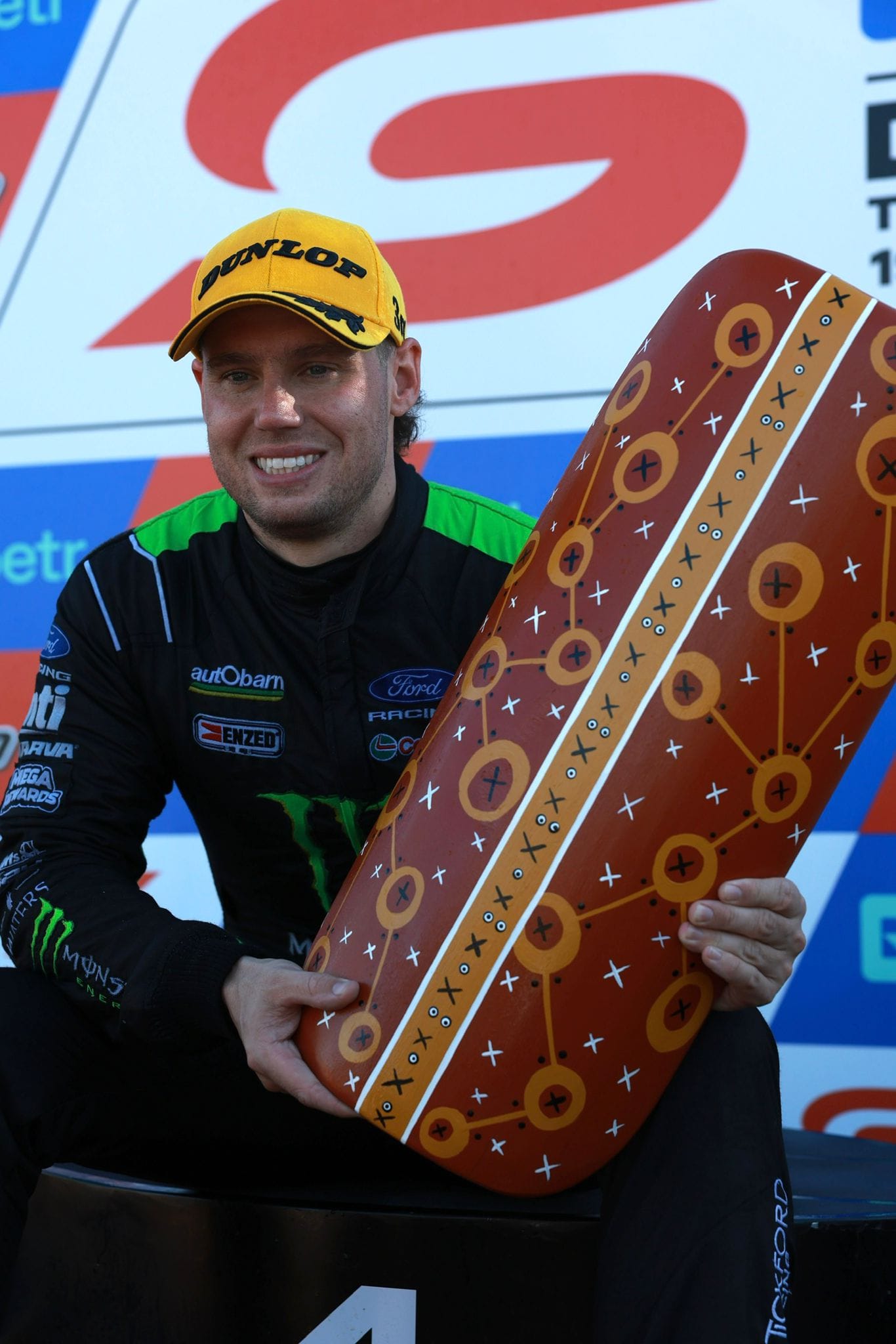 A Supercars driver in a yellow cap holds an Indigenous Round trophy piece on the Darwin Triple Crown podium.