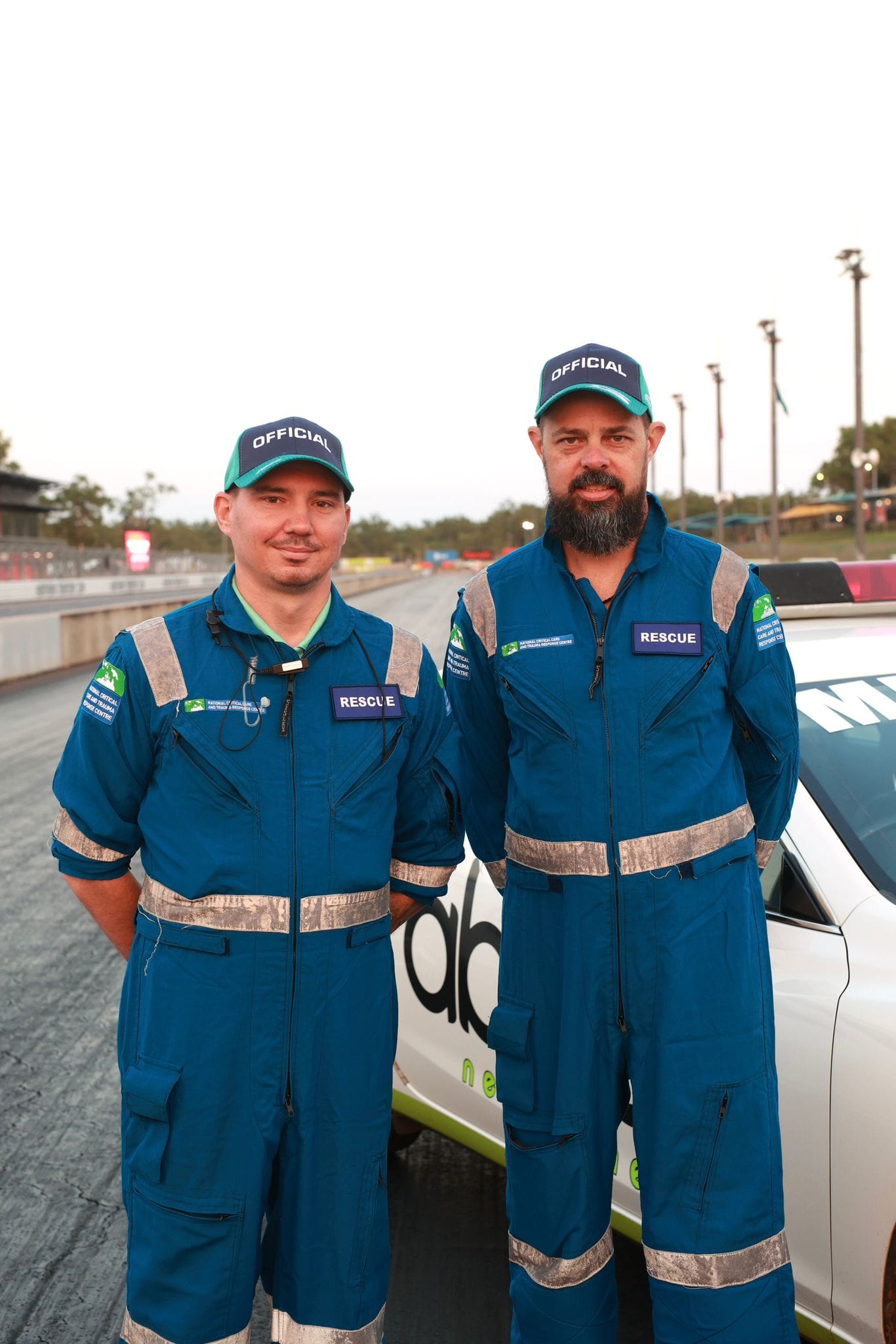 Two rescue crew members in blue uniforms pose beside an official vehicle on the Hidden Valley Raceway main straight.