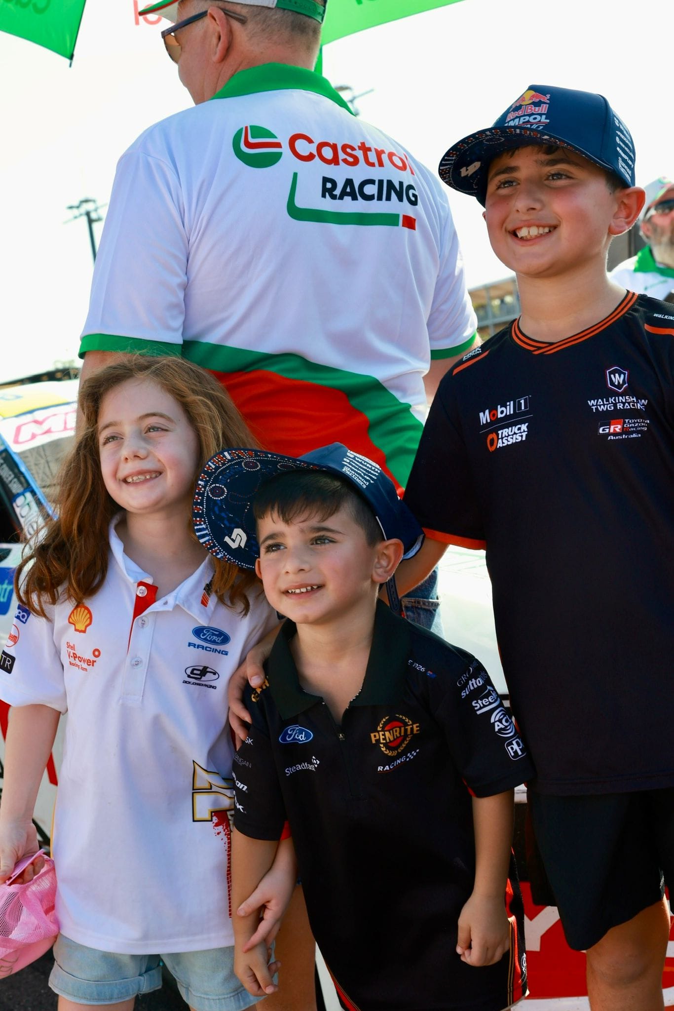 Three young motorsport fans smile beside a race car during the Darwin Supercars grid walk.