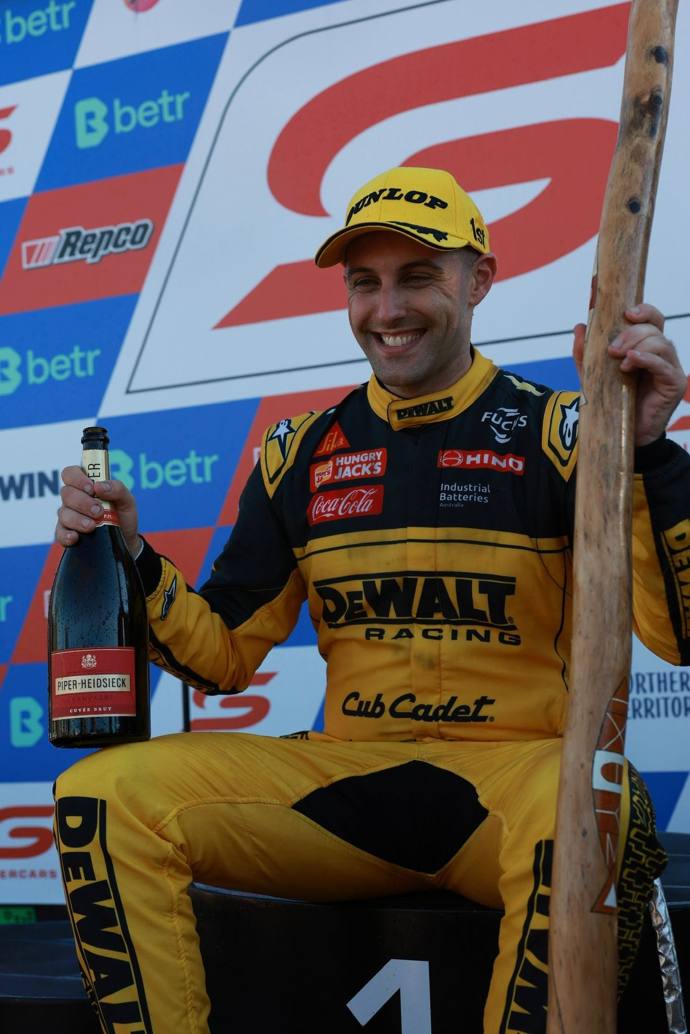 The race winner smiles on the Darwin Triple Crown podium while holding a champagne bottle and carved trophy piece.