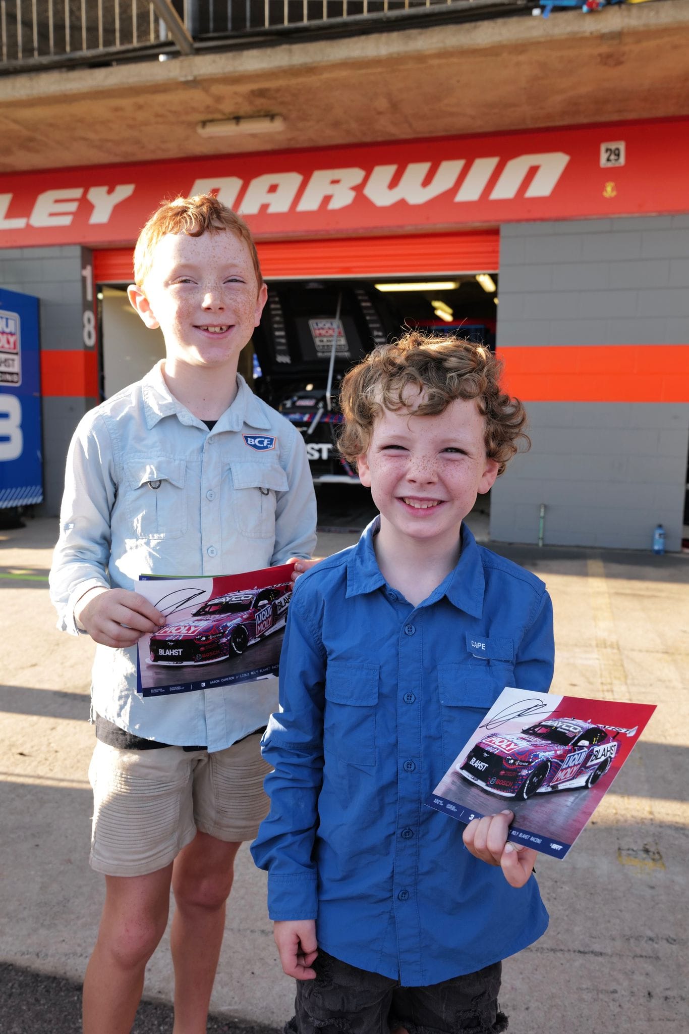 Two young fans smile while holding signed Supercars posters in front of the Hidden Valley Darwin pit garages.