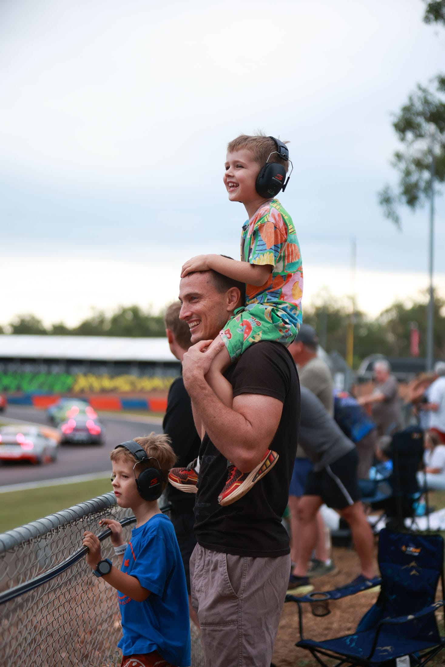 An adult and two children wearing ear protection watching Supercars race from the spectator area at Hidden Valley Raceway.