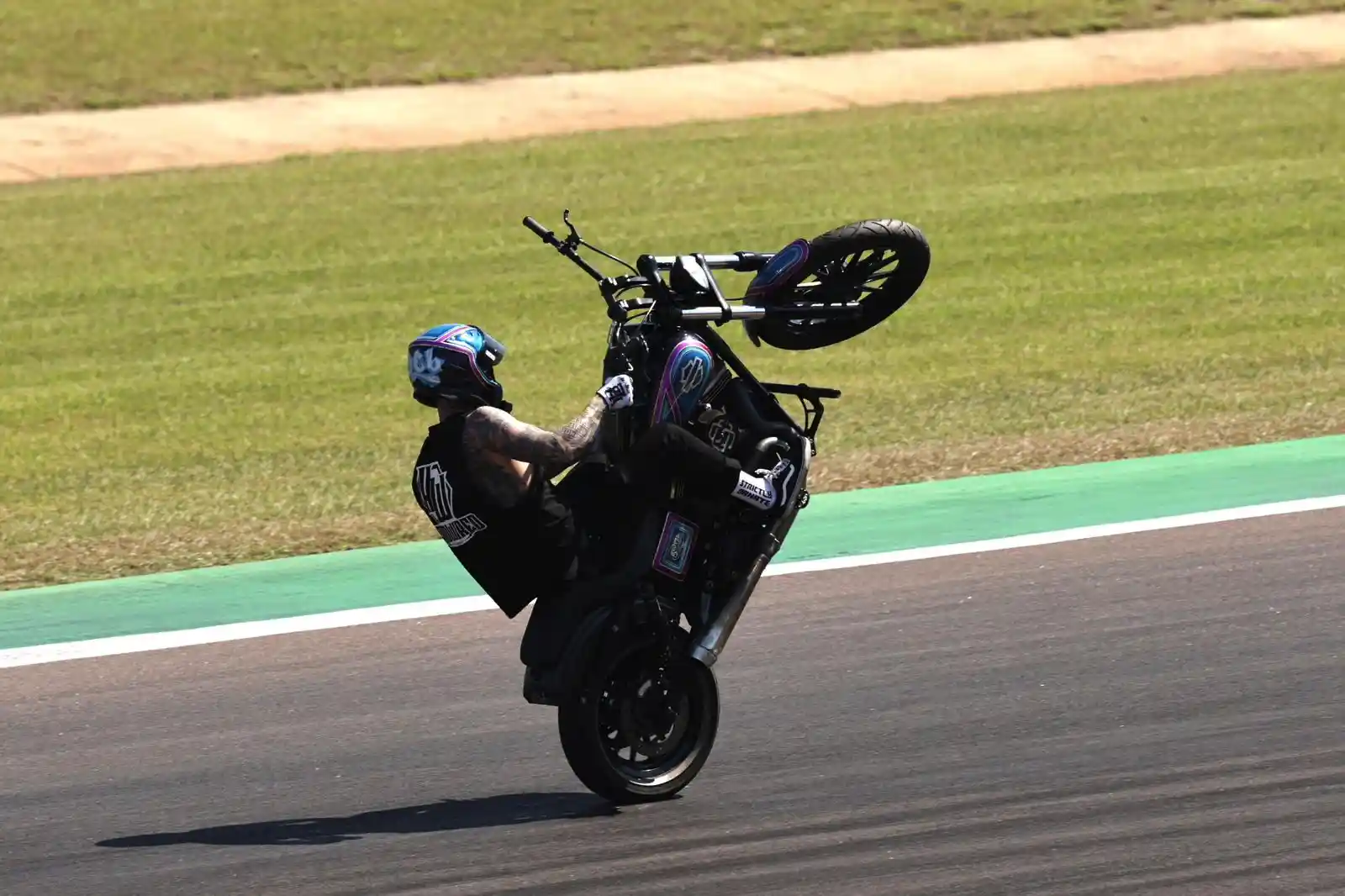 Motorcyclist performing a wheelie on a track, wearing a blue helmet and black attire, with grass and asphalt in the background.