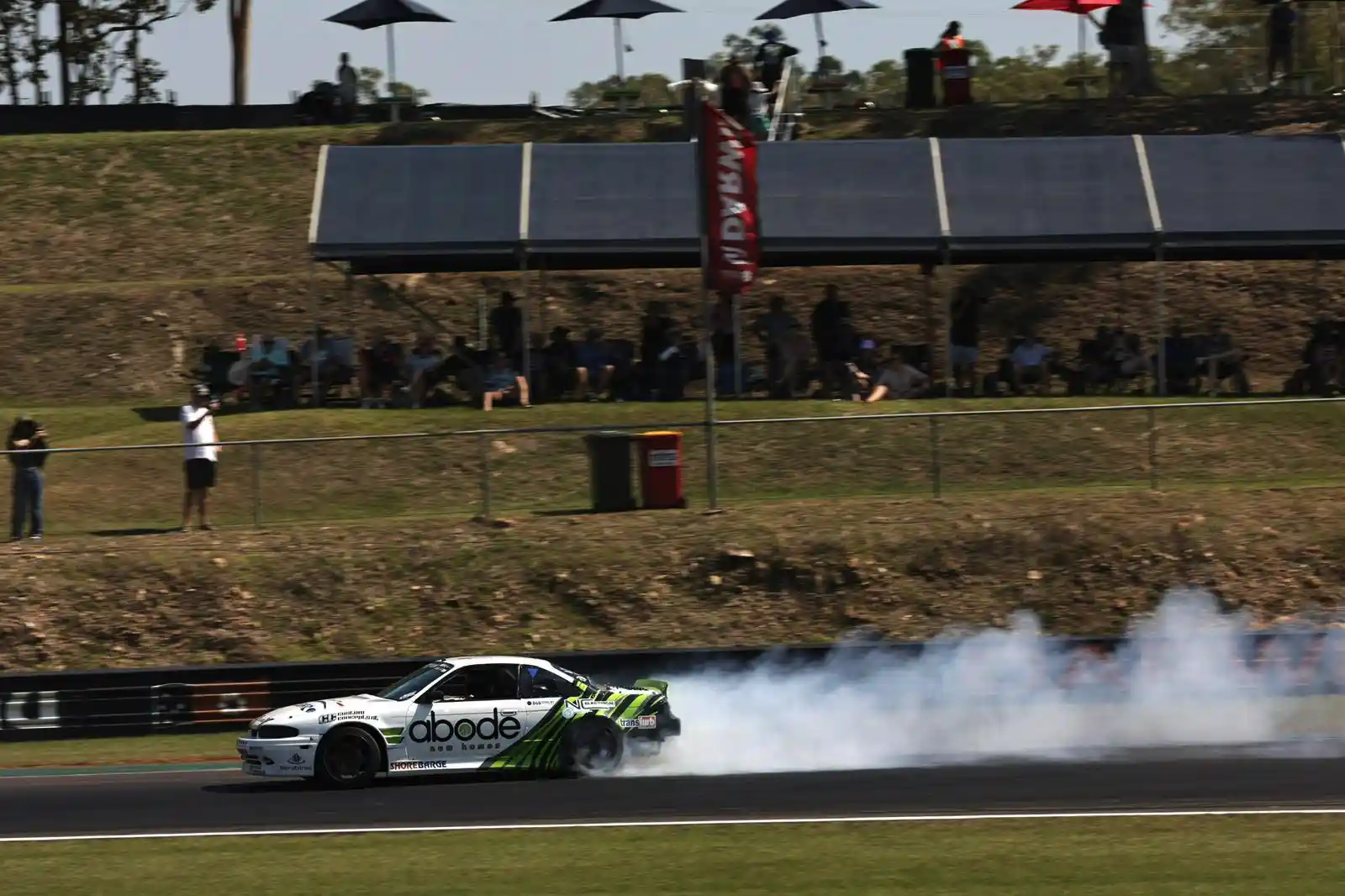 A white and green car drifts on a racetrack, producing smoke, with spectators watching from a grassy hill under umbrellas.