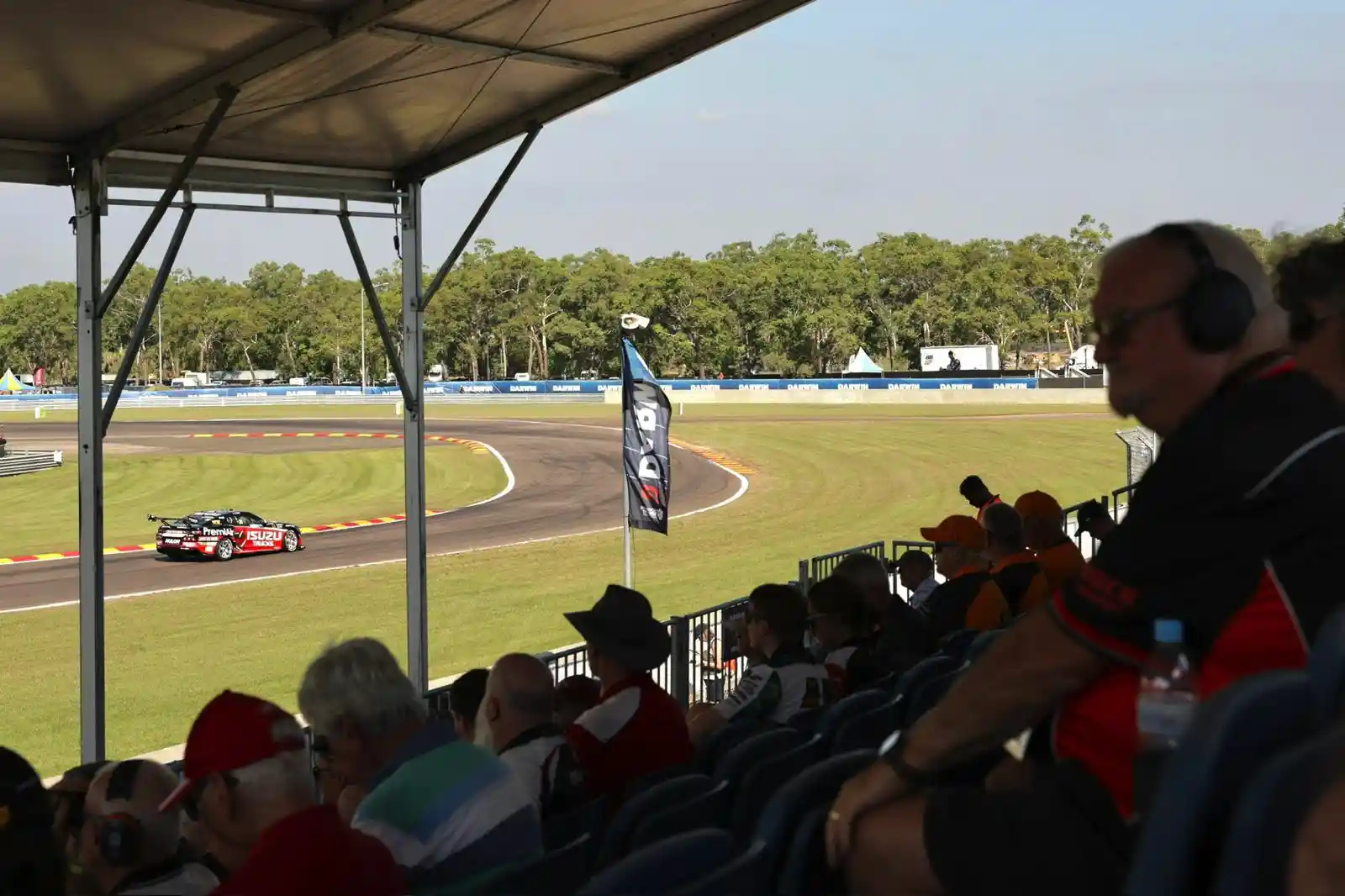 Spectators watch a car race from shaded stands, with a race car speeding on the track and trees in the background.