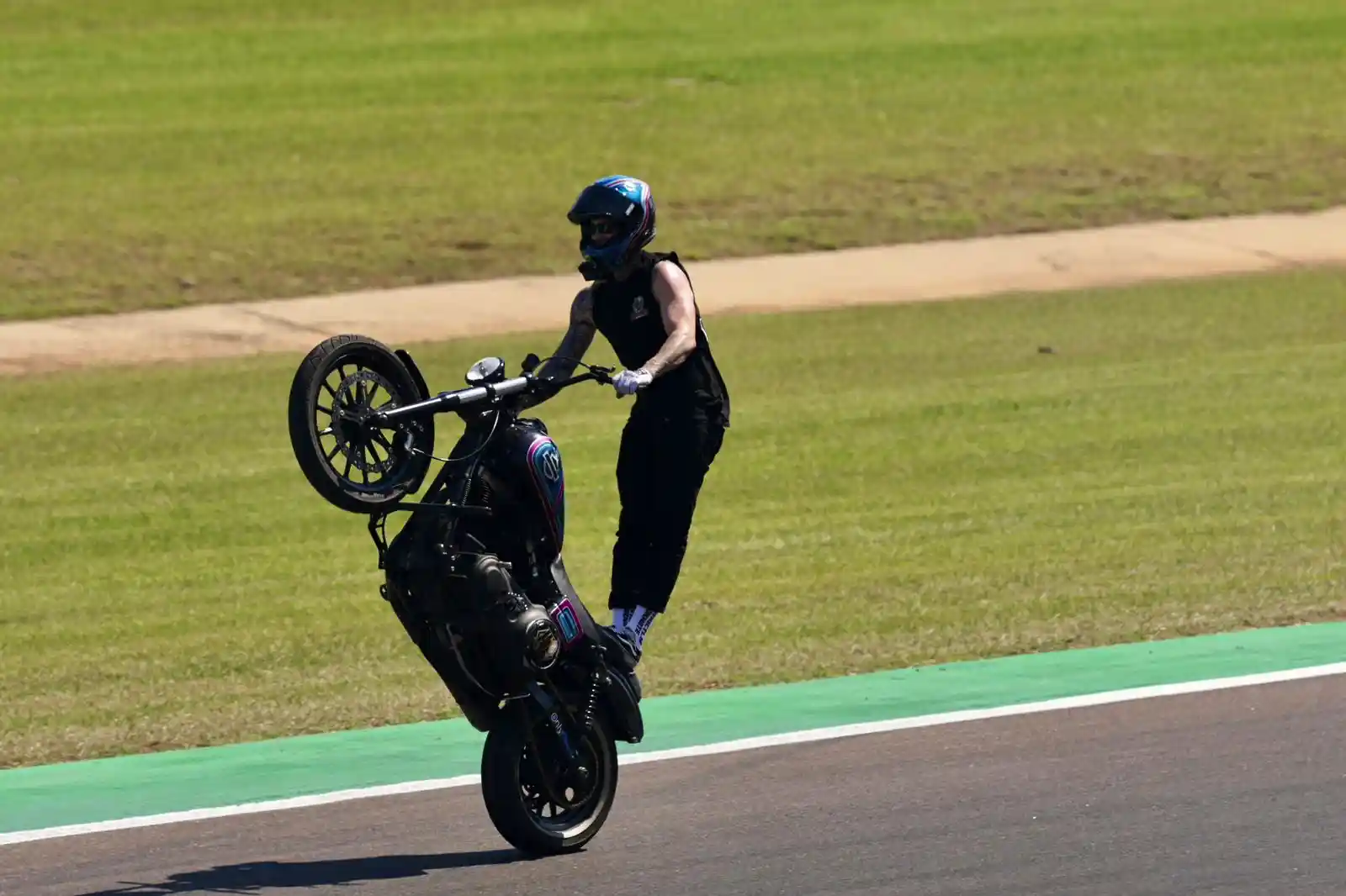 Motorcyclist performing a wheelie on a racetrack, wearing a helmet and black attire, with grassy areas in the background.