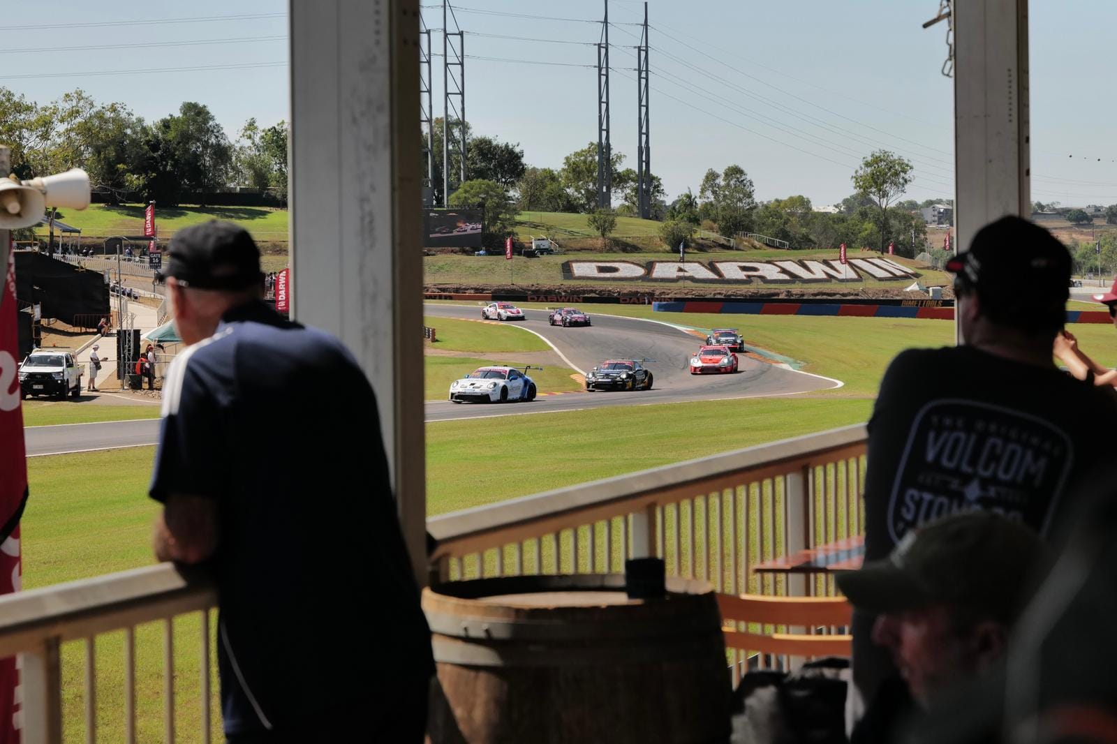Spectators watch Supercars race around a corner at Hidden Valley Raceway from a shaded balcony overlooking the circuit.
