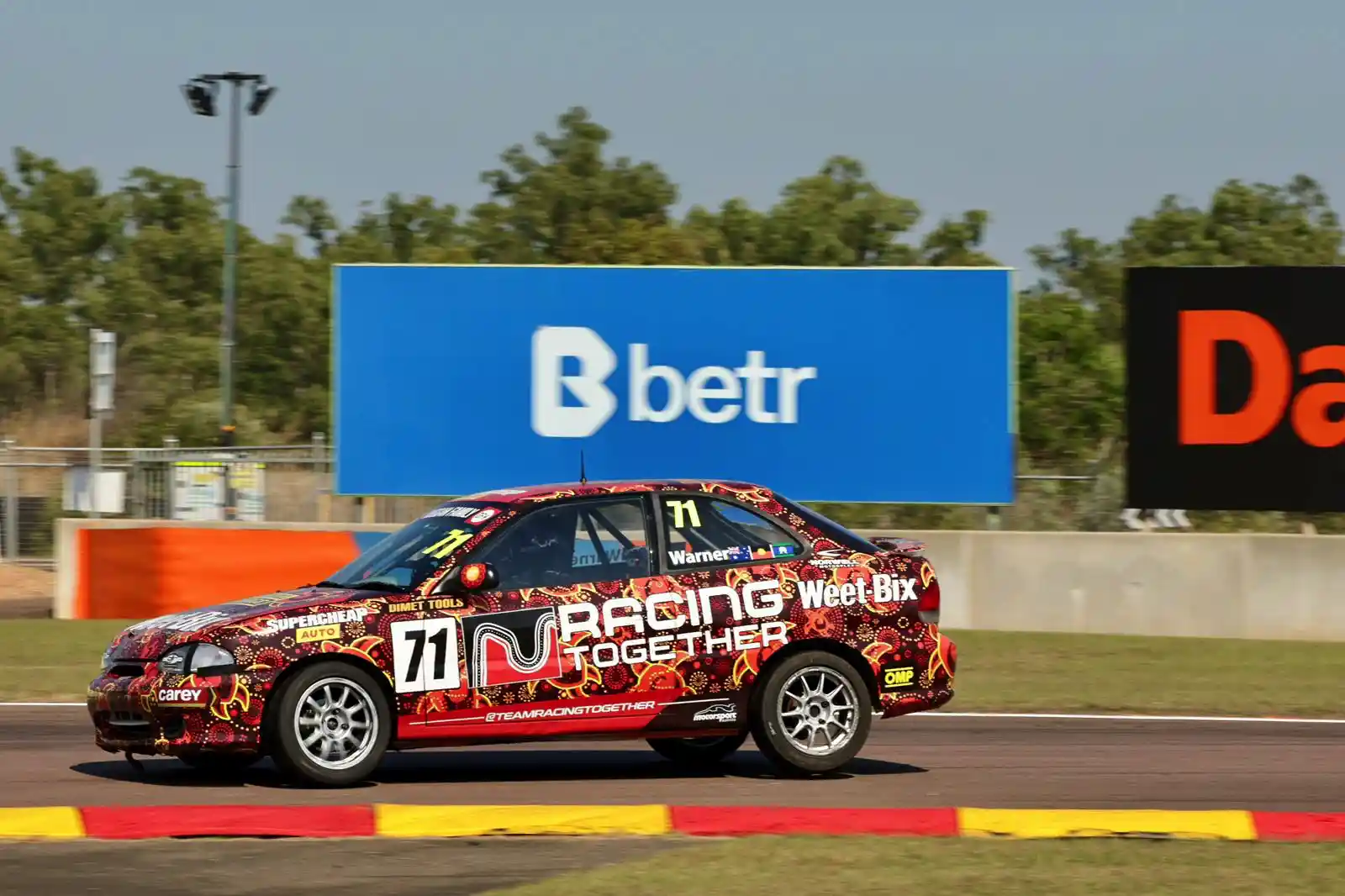 Red racing car with number 71, featuring "Racing Together" and sponsor logos, speeding on a track with trees and advertisements in the background.
