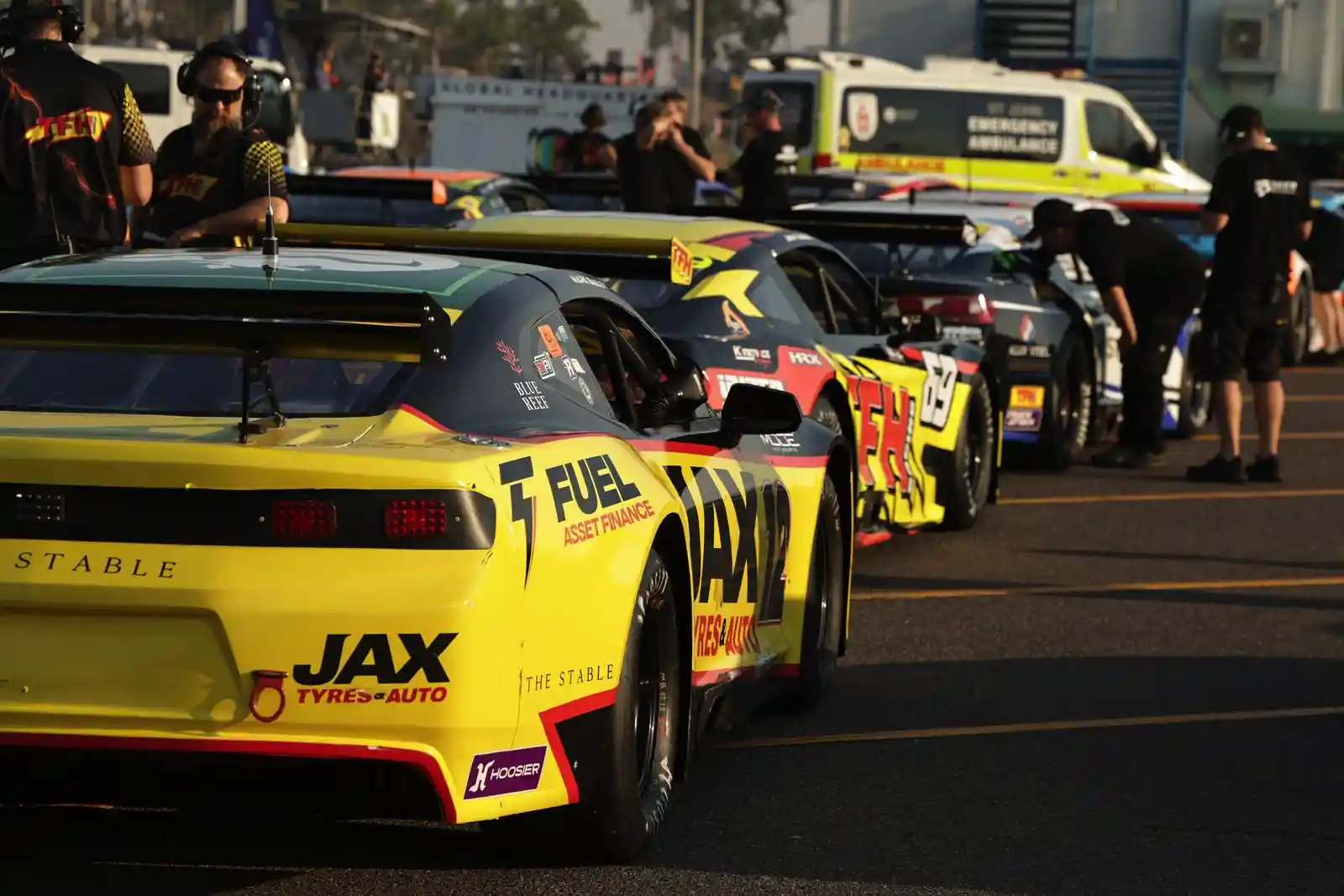 Race cars lined up on a track with drivers and crew nearby, featuring prominent yellow and black designs, and an ambulance in the background.