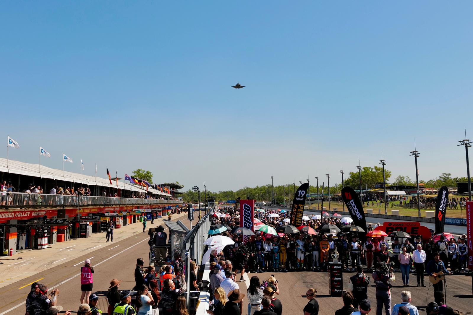 A wide view of the Darwin Triple Crown pre-race ceremony shows teams, officials and spectators gathered on the grid as a jet flies overhead.
