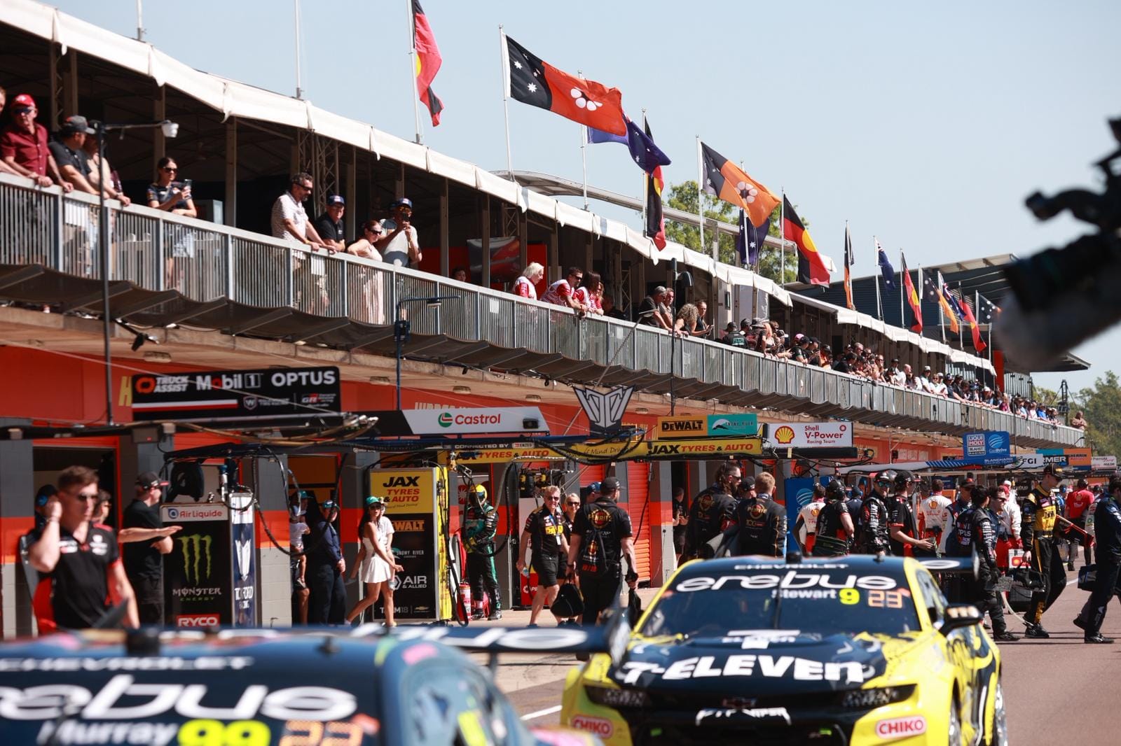 Supercars sit in pit lane at Hidden Valley Raceway as crews and spectators watch from the garages and balcony above.