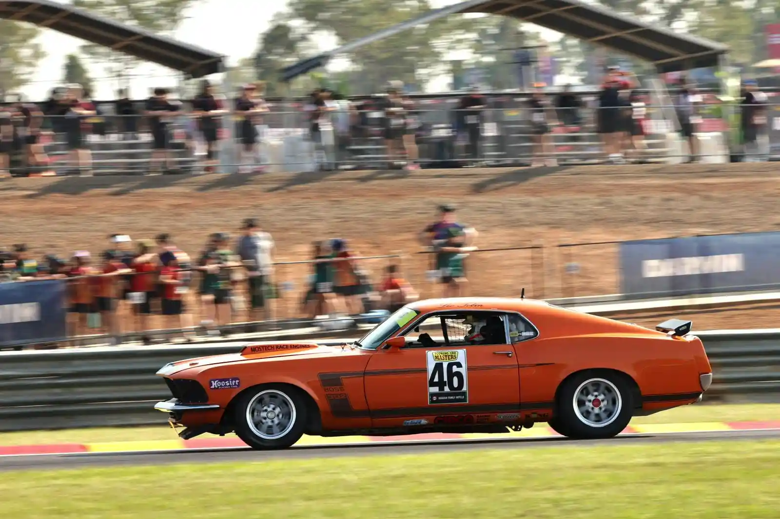 An orange classic race car with the number 46 speeds on a track, with spectators in the background.
