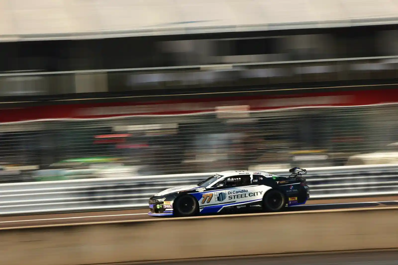 A race car speeds along a track, with motion blur emphasizing its velocity. The car is branded with "Steel City" and number 70.
