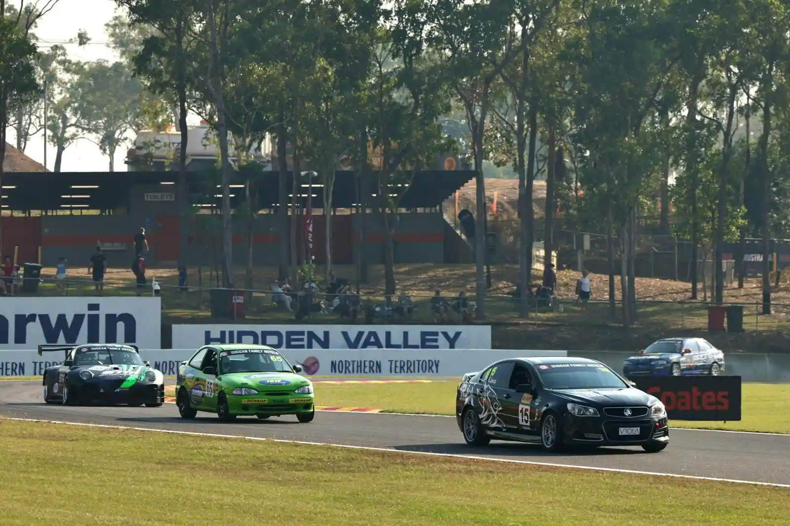 Cars racing on a track at Hidden Valley, surrounded by trees and spectators, with banners and a clear sky in the background.