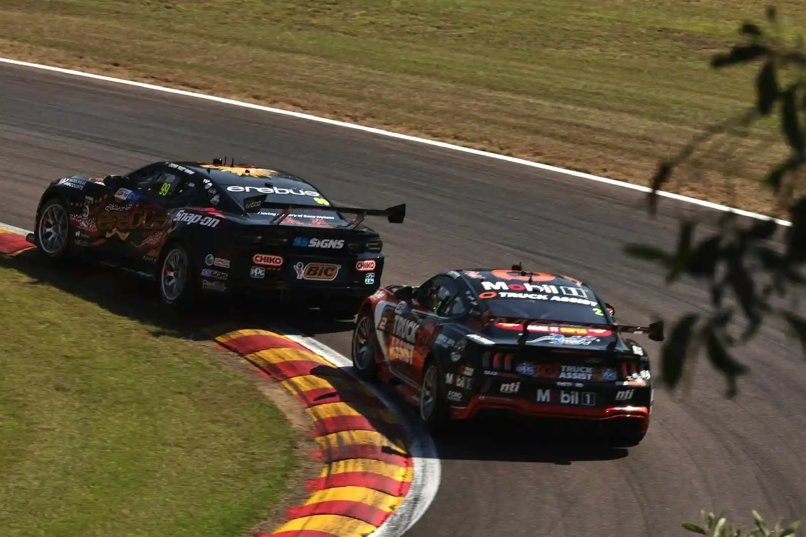 Two race cars with sponsor logos speed around a curved track, surrounded by grass and a few trees.