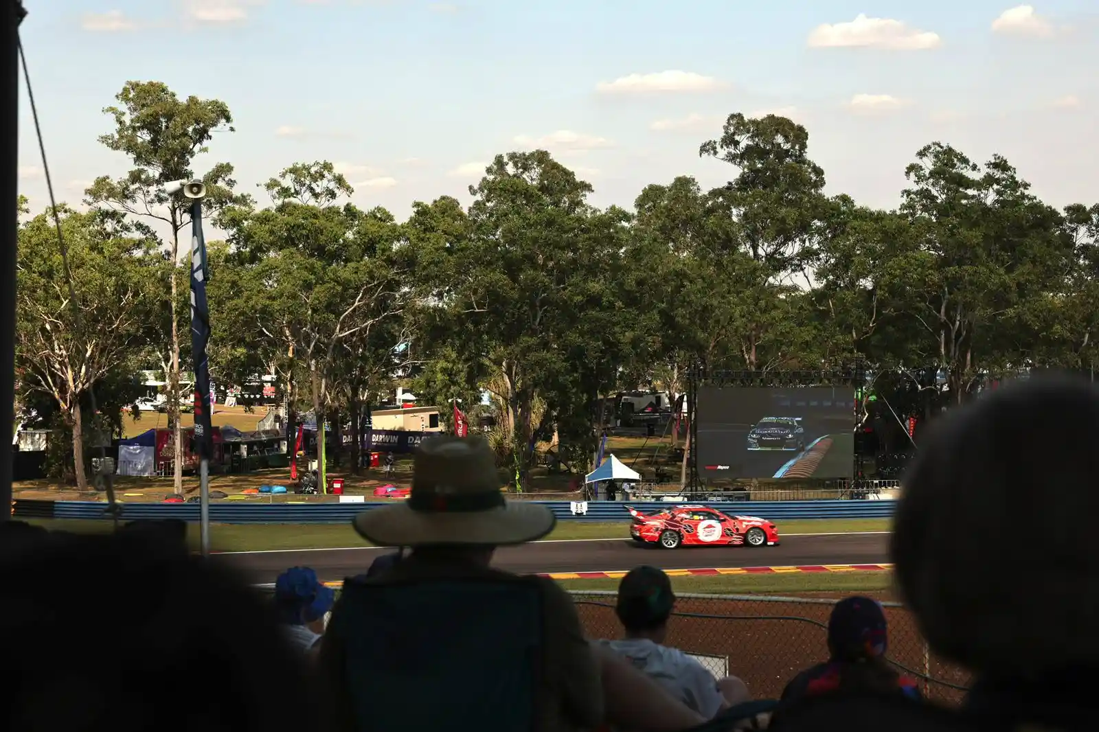 Spectators watch a red race car on a track surrounded by trees, with a large screen displaying the race in the background.