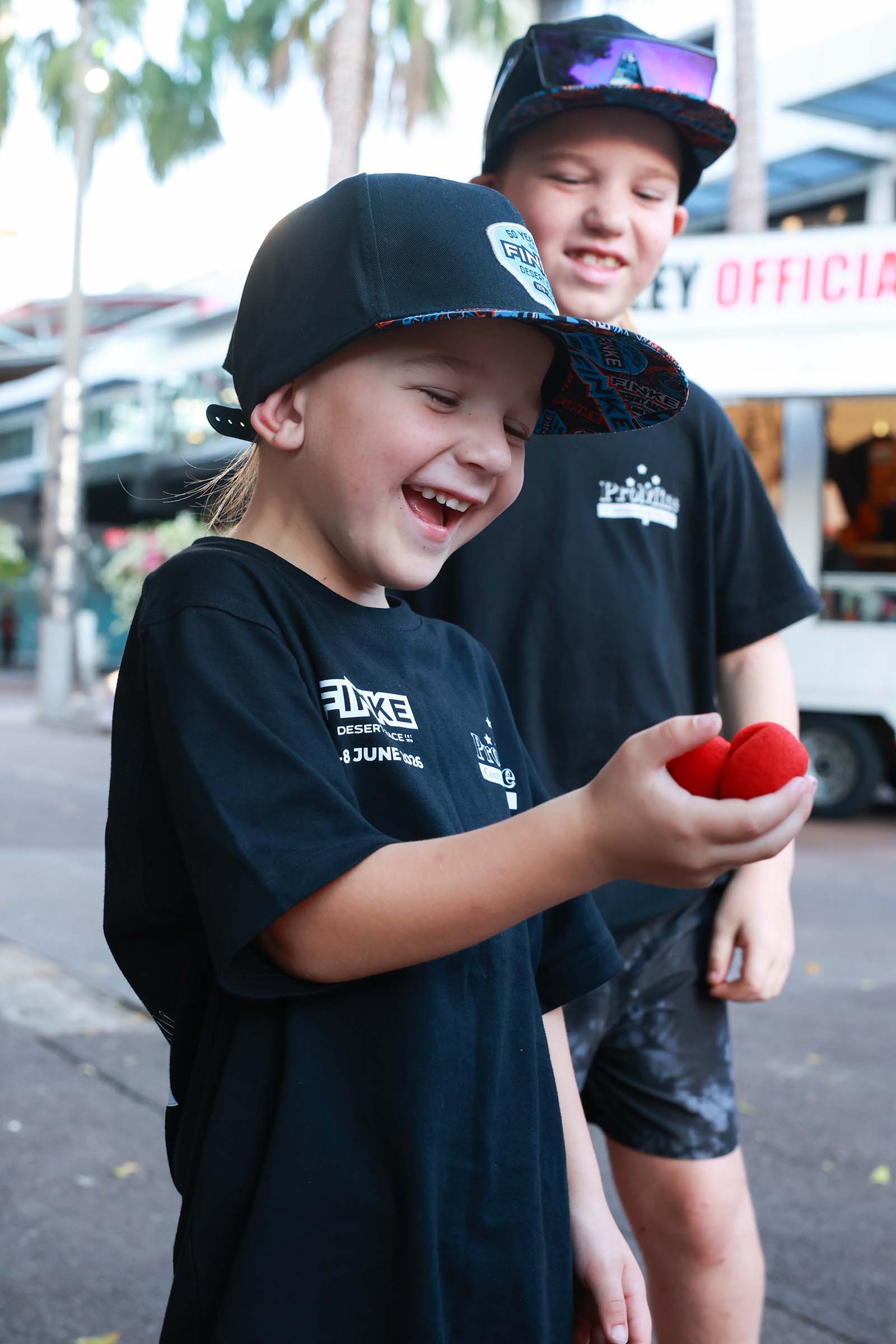 Two children wearing black Finke Desert Race caps watch a street performer with red sponge balls.