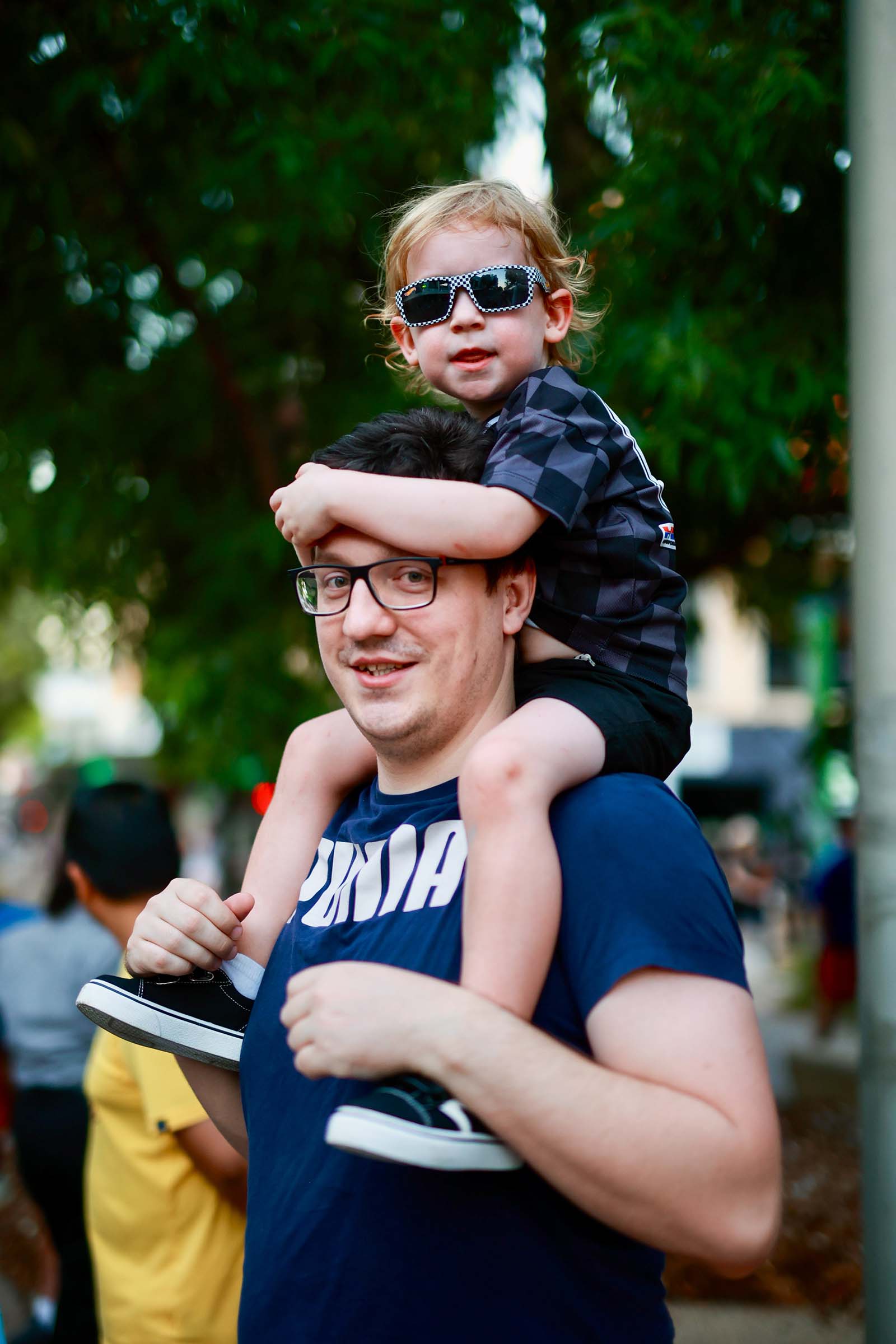 A child wearing sunglasses sits on an adult’s shoulders while watching the Darwin Supercars street parade.