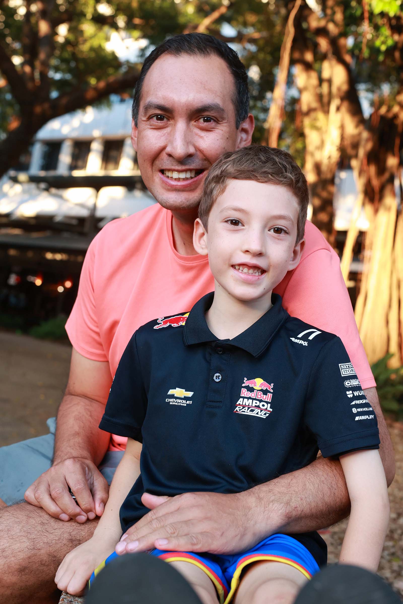 An adult and child smile for a photo, with the child wearing a Red Bull Ampol Racing shirt.