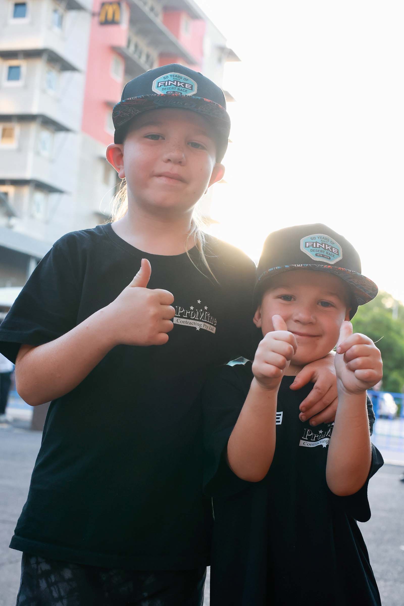 Two children wearing Finke Desert Race caps give thumbs up while standing together in Darwin city.