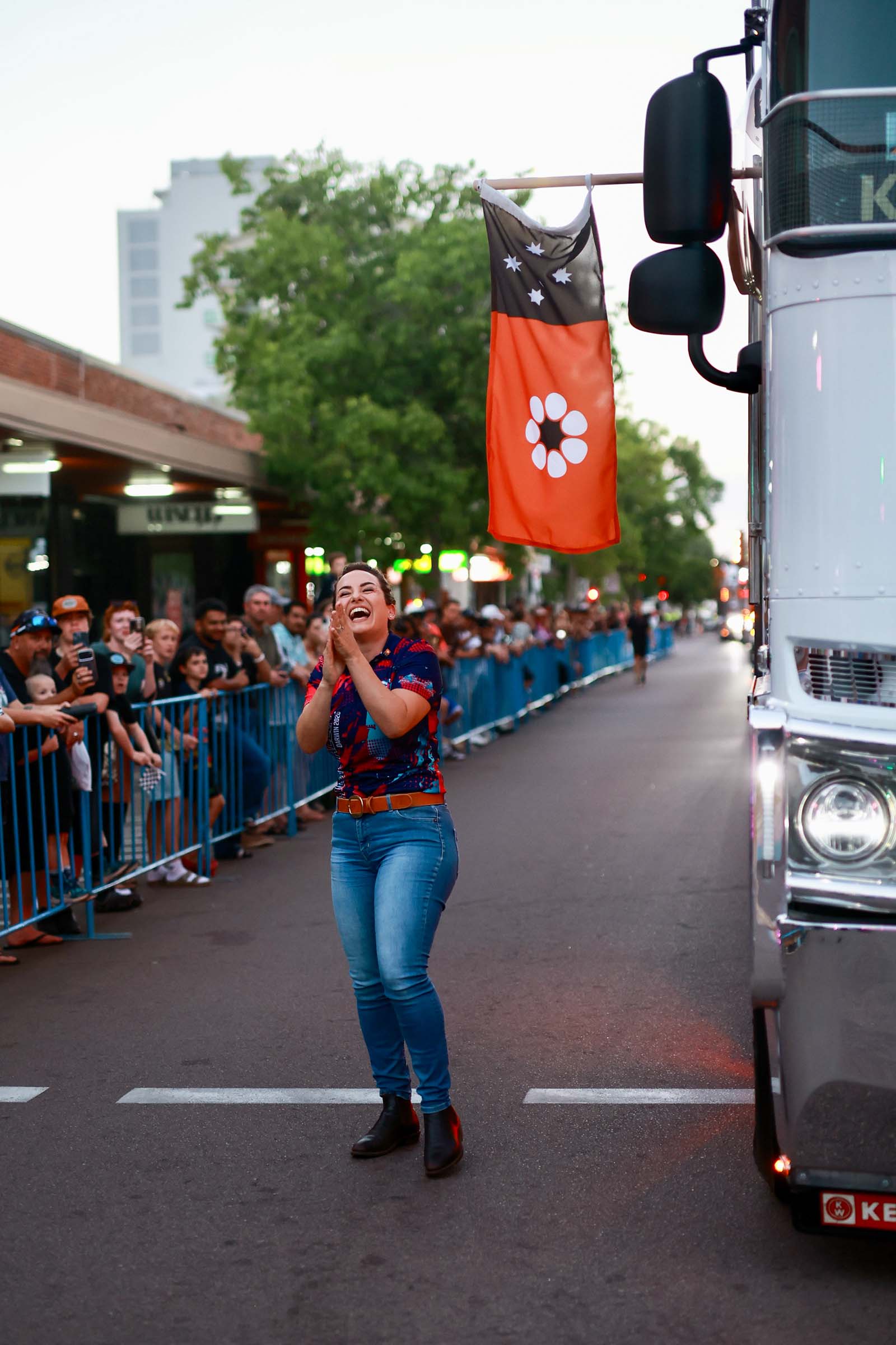 A woman clapping beside a Supercars truck carrying a flag during the Darwin street parade, with crowds lining the road.