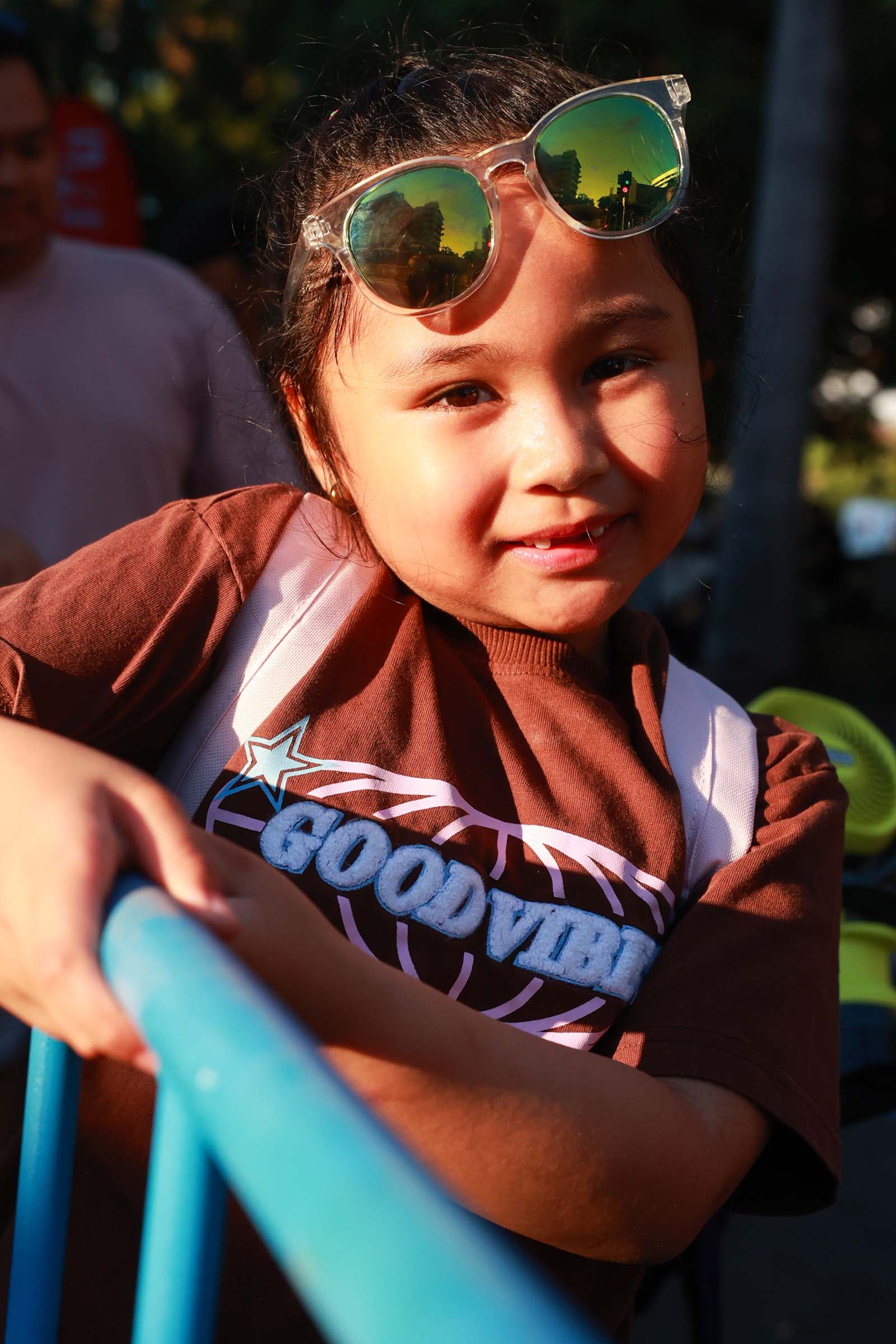 A child wearing sunglasses on her head leans against a blue crowd barrier in warm afternoon light.
