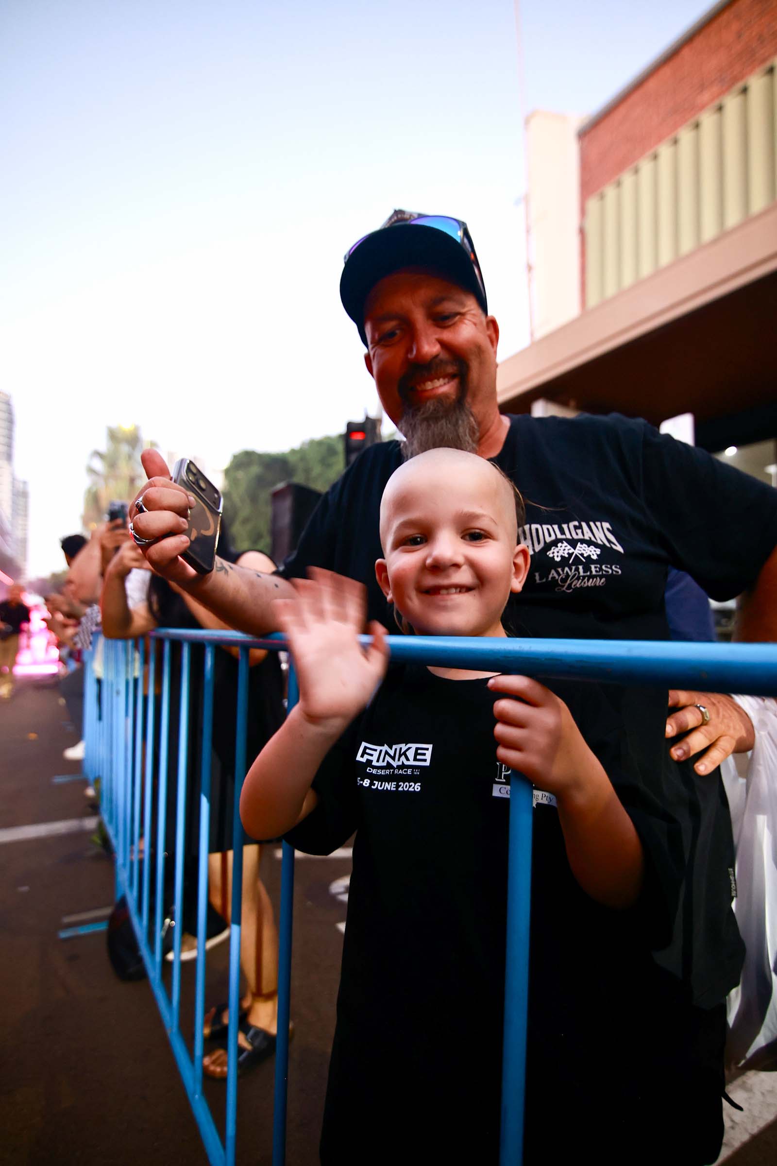 A child waves from behind a crowd barrier while watching the Darwin Supercars street parade with an adult.