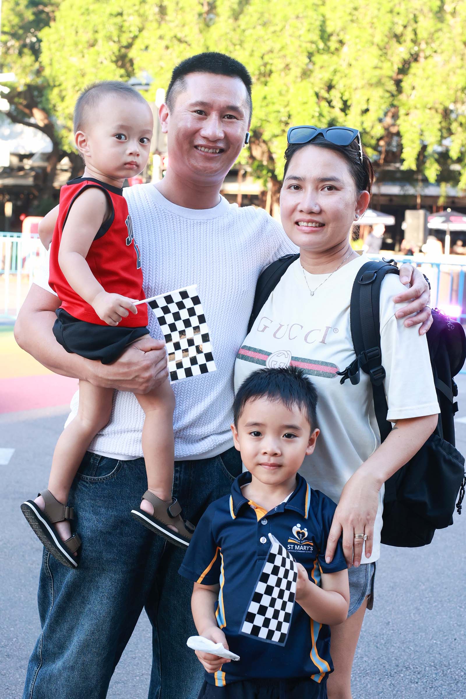 A group of spectators pose together at the Darwin Supercars street parade, with children holding chequered flags.