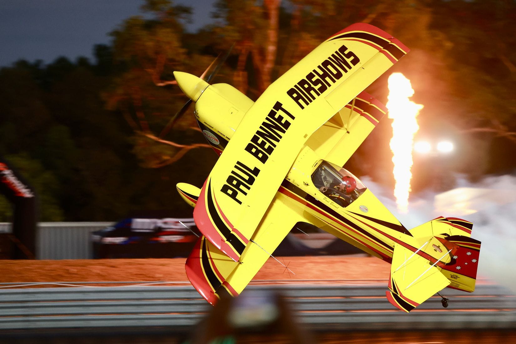A Paul Bennet Airshows plane performs low over the track with flames and smoke behind it during a motorsport display.
