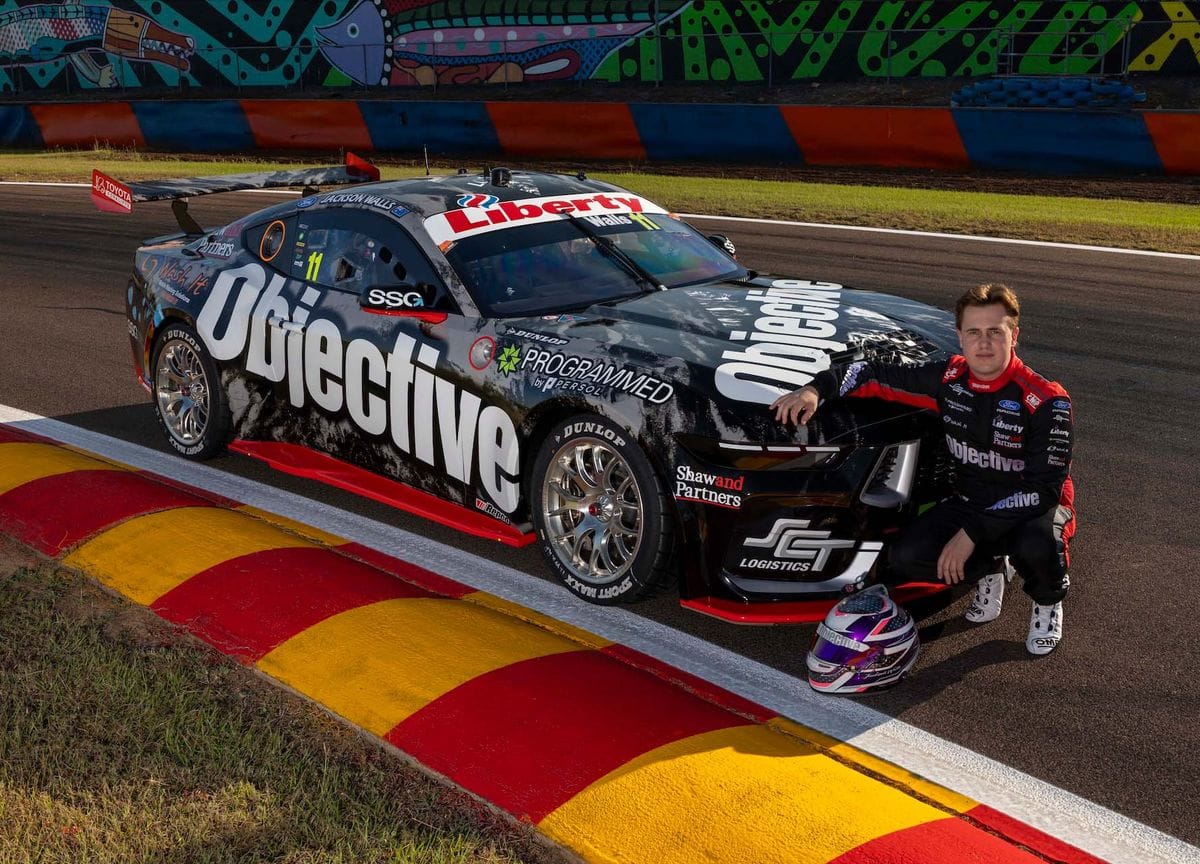 Driver standing beside an Objective Racing Supercar on track, with a racing helmet placed near the front wheel.
