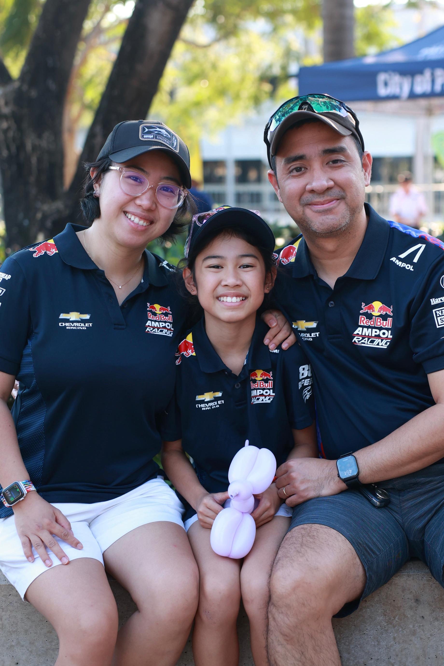 A smiling family of three in matching navy shirts and caps, sitting outdoors. The child holds a white balloon animal. Trees are in the background.