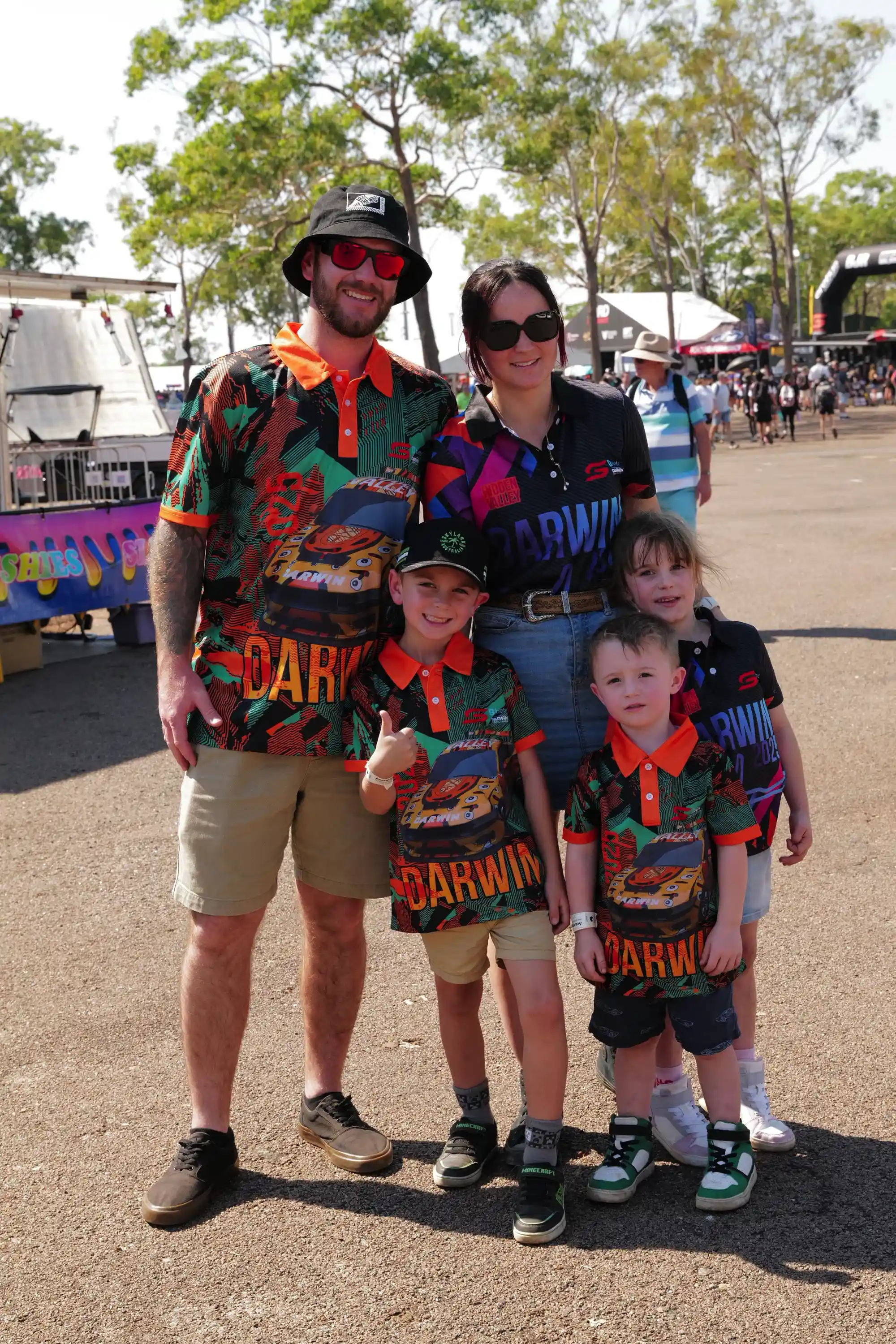 Family wearing matching "Darwin" shirts and hats, posing outdoors at an event. Trees and tents visible in the background.
