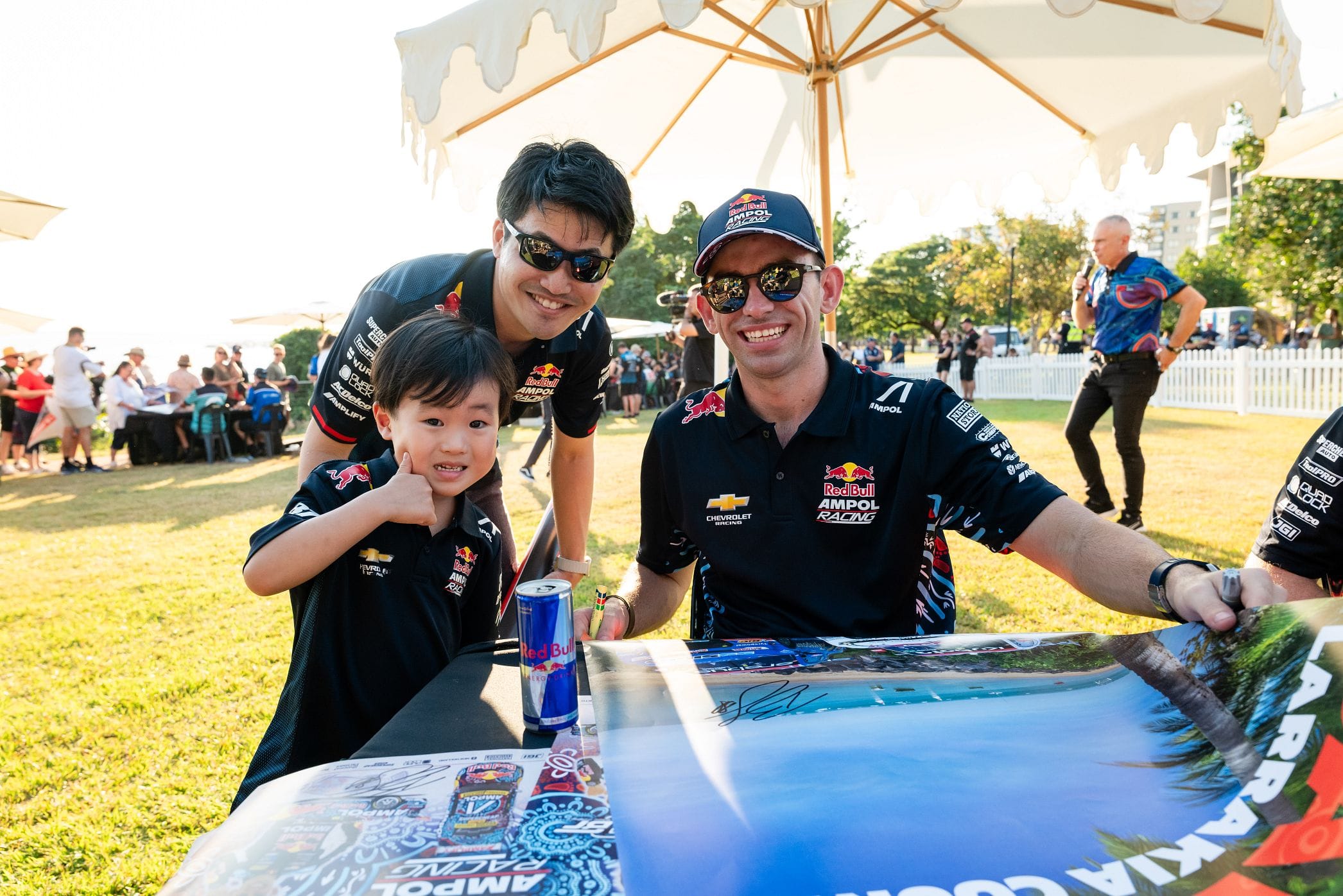 Two men in Red Bull Racing polos pose with a young boy at an autograph table under a large white umbrella; crowd and a speaker in the background.