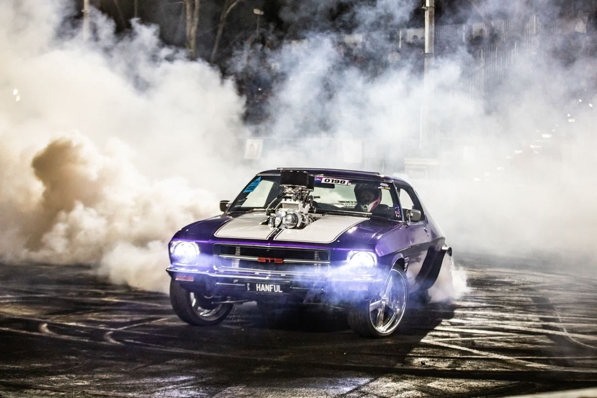 Purple muscle car with exposed engine performing a burnout, engulfed in thick white smoke, bright headlights cutting through the haze.