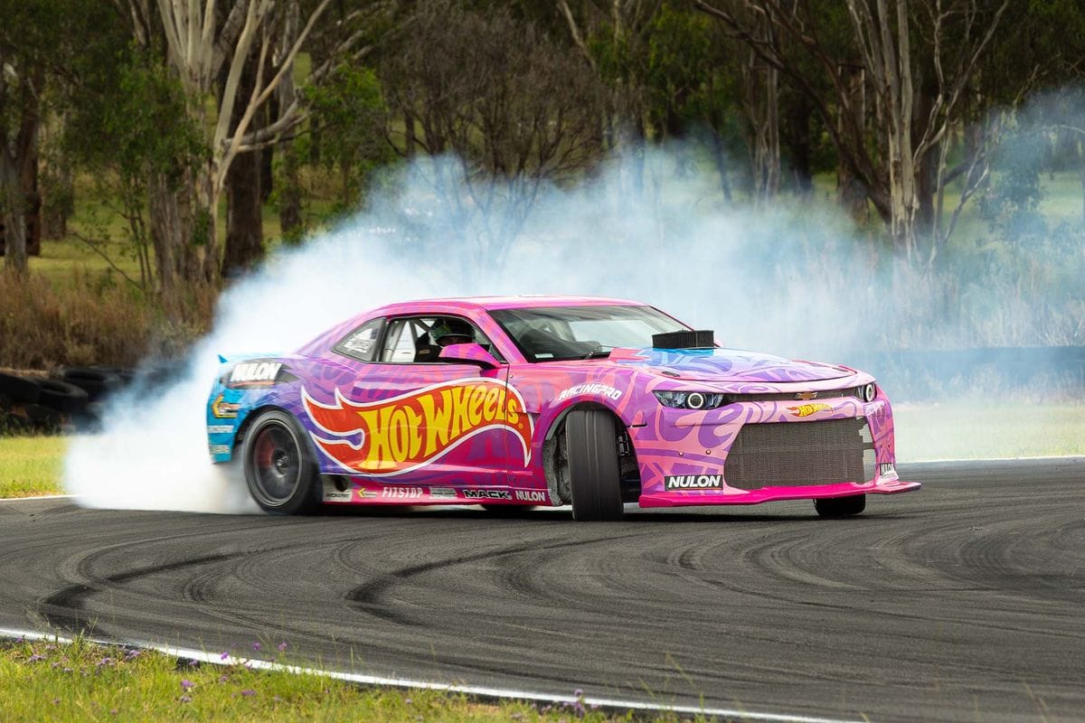 A pink car on track doing a burnout with smoke coming from the back wheels and "Hot Wheels" text on the side of the car
